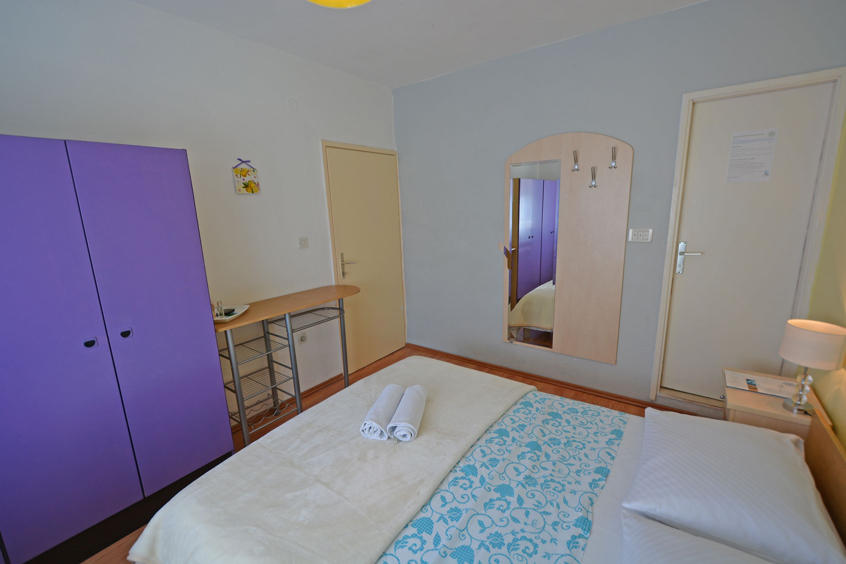 A hotel room with a bed, a desk, a purple wardrobe, and a mirror.