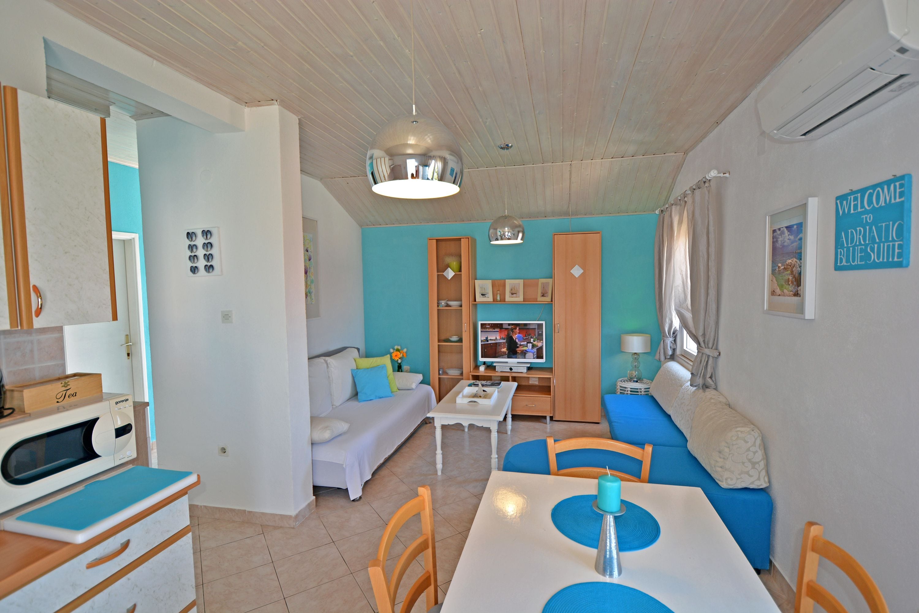 A modern living room with a sofa, dining table, and TV. There is a kitchen area with a microwave and cabinets. The room has a wooden ceiling and a sign welcoming guests to the suite.