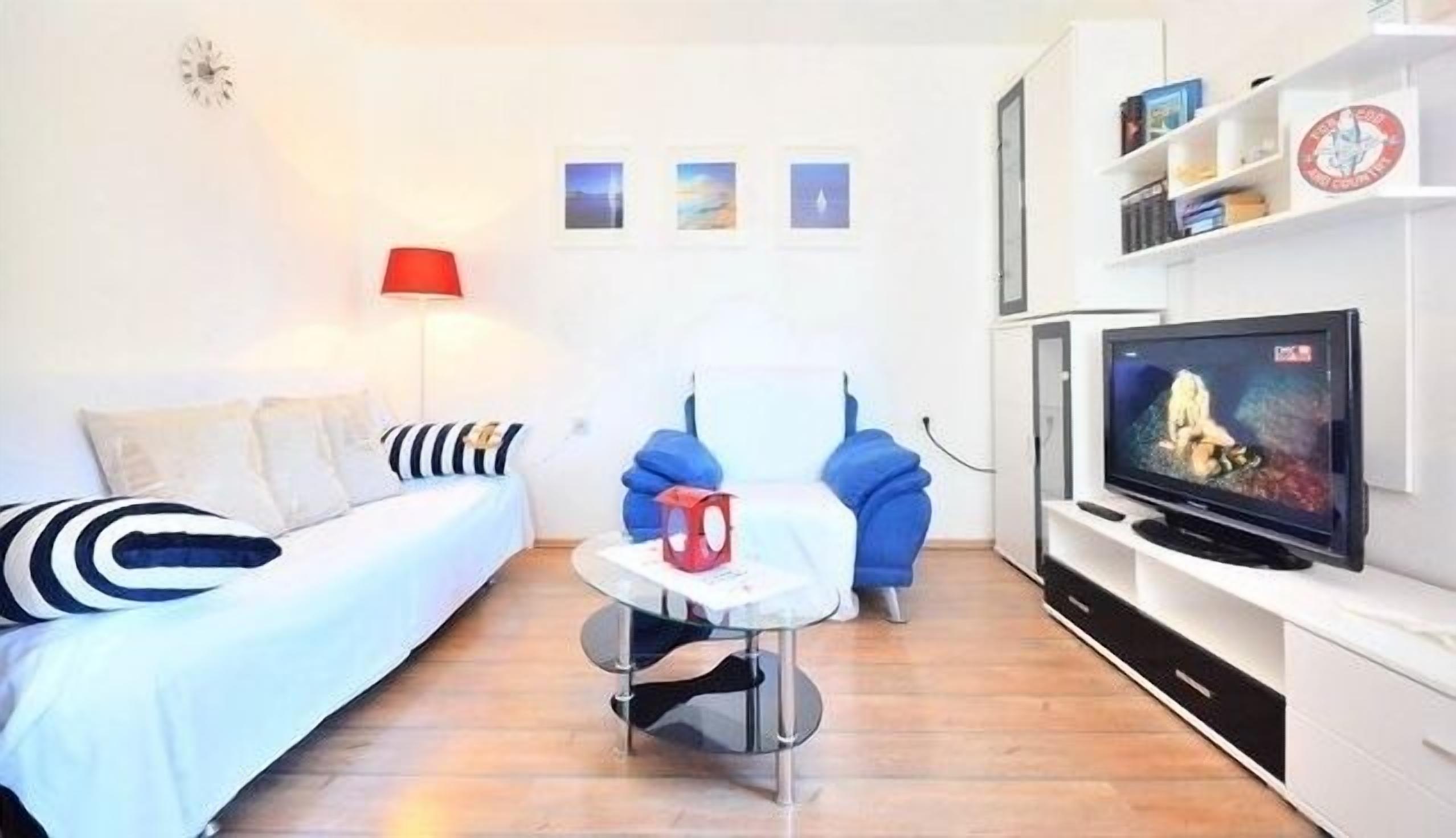 A modern living room with a sofa, a red lamp, a small table, a blue chair, and a TV on a white cabinet.