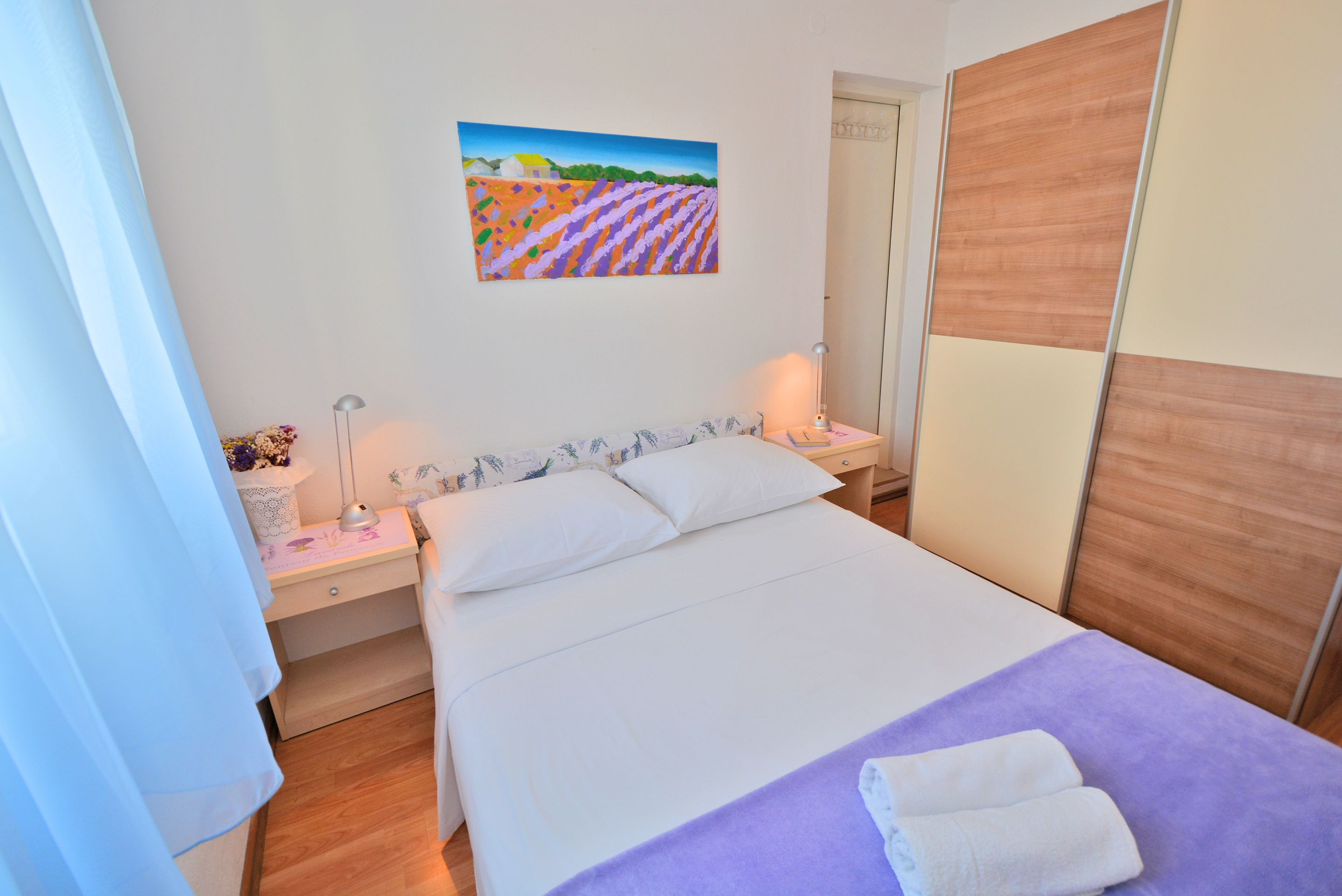 A neatly arranged hotel room with a large bed, a bedside table with a lamp, a nightstand, and a painting on the wall.
