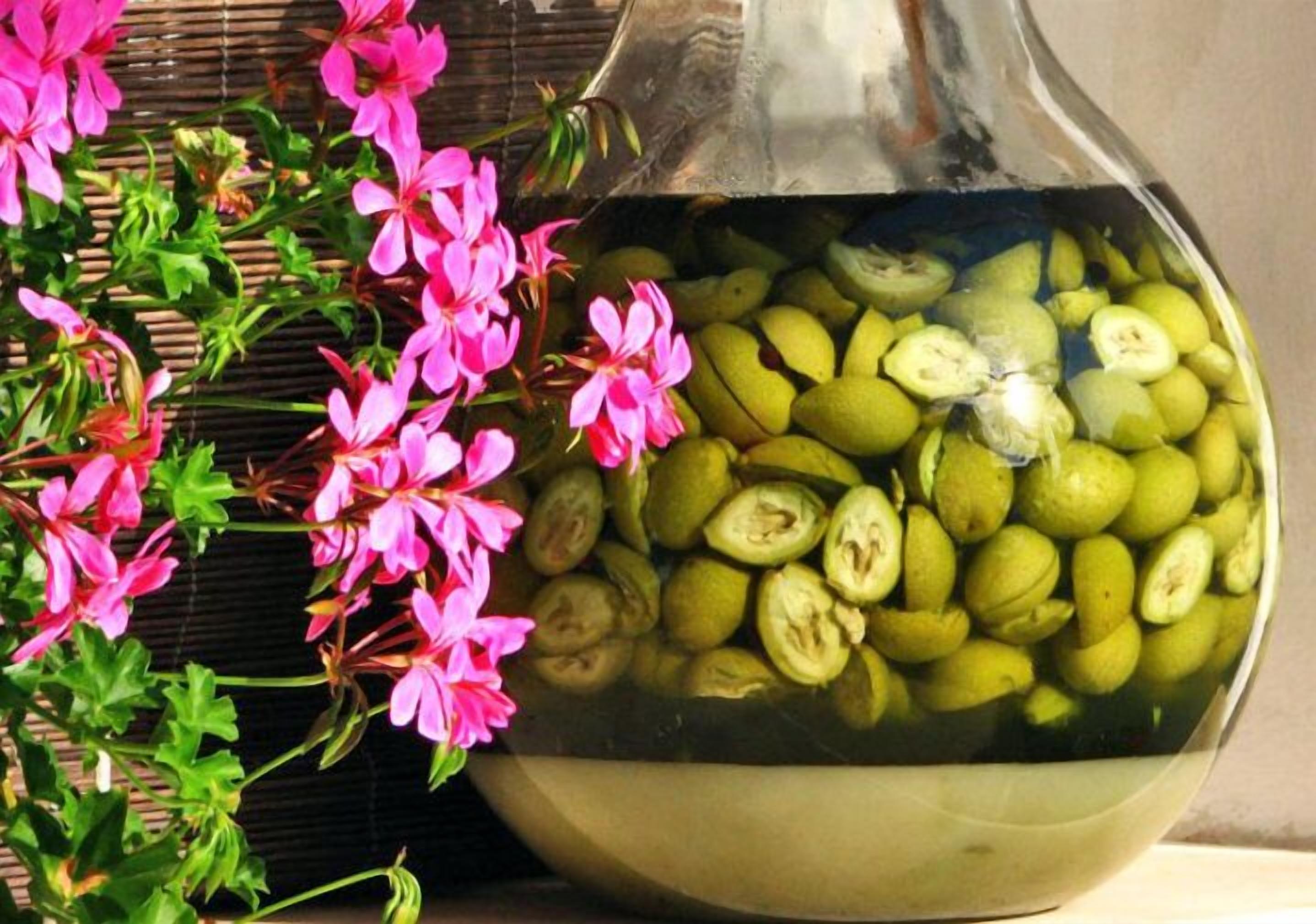 A bowl filled with sliced green fruits, possibly lemons, next to a plant with pink flowers.
