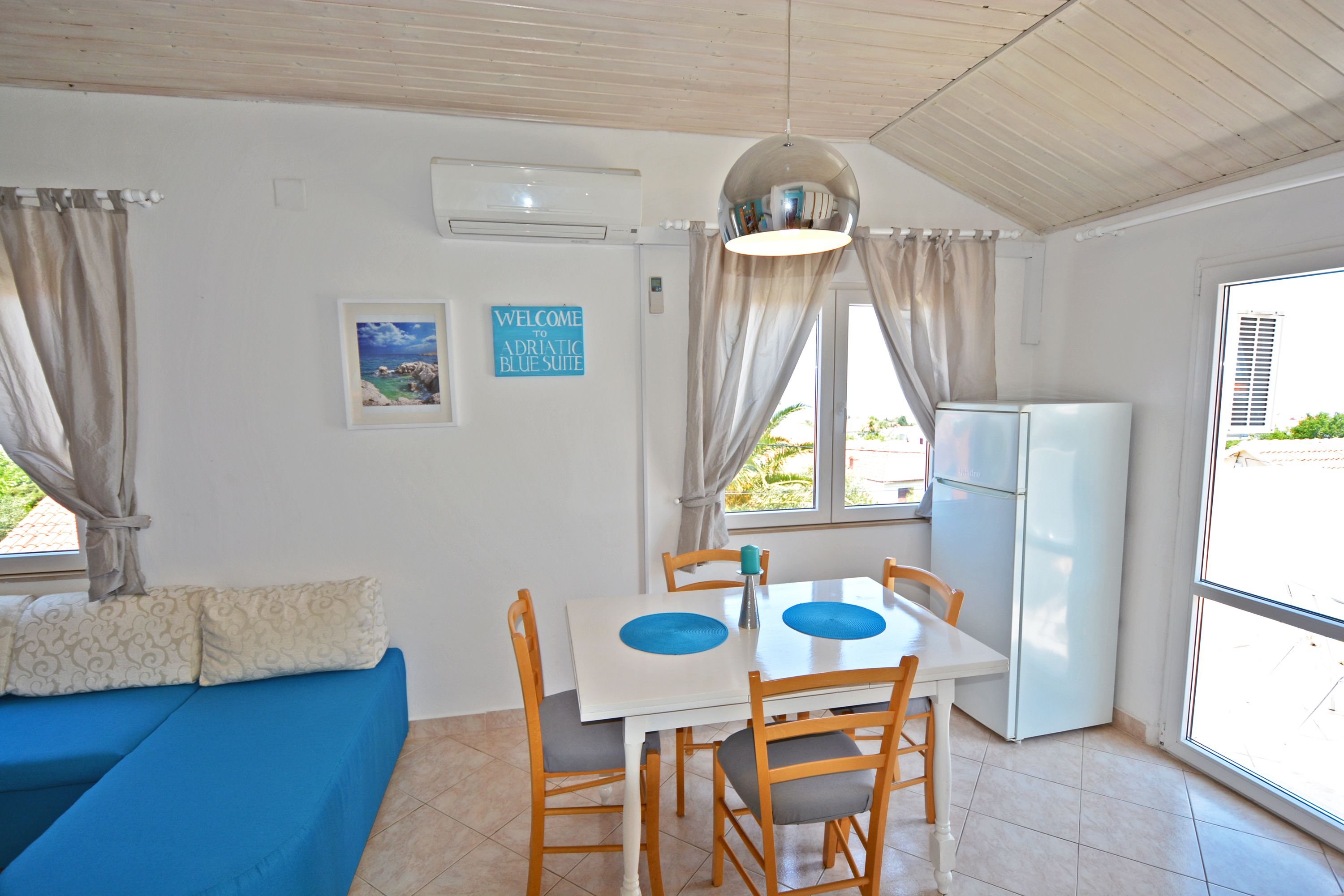 A room with a blue sofa, a dining table with chairs, a refrigerator, and a window with curtains.