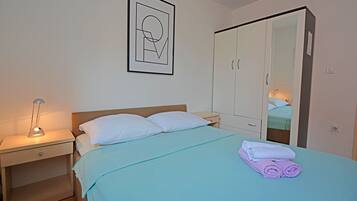 Una cama bien tendida con colcha azul, almohadas blancas y toallas enrolladas. Se observa una mesita de noche con una lámpara y un cuadro enmarcado en la pared.
