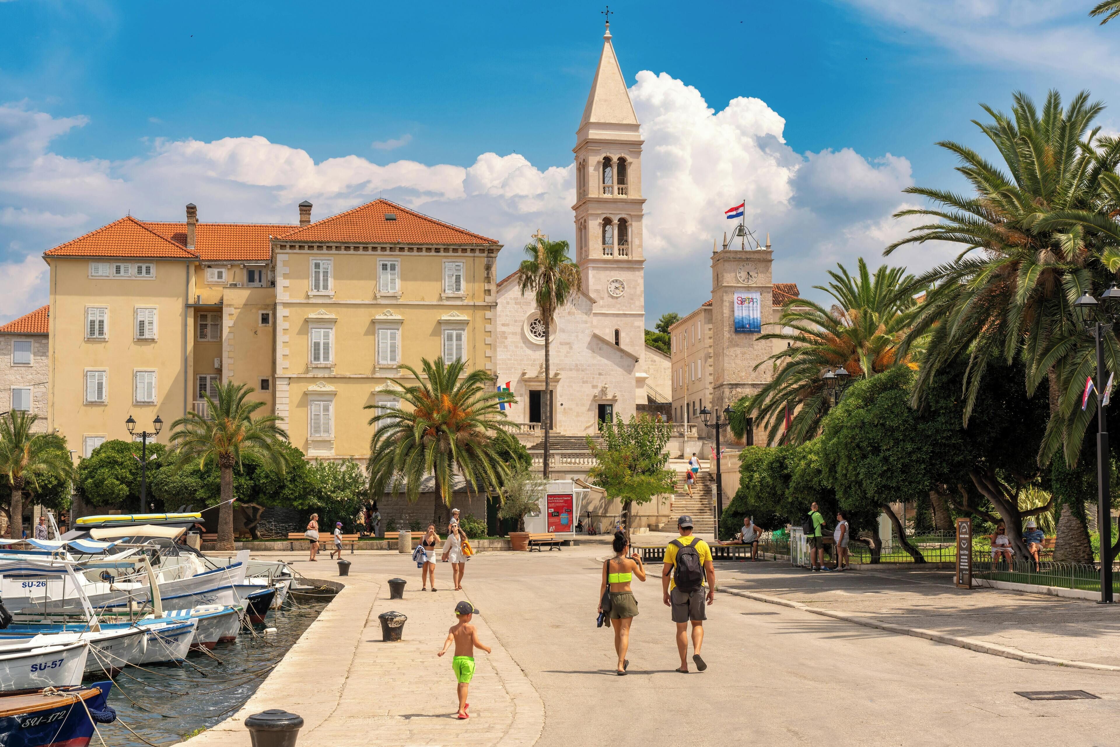 A waterfront promenade with buildings, a church tower, and palm trees.