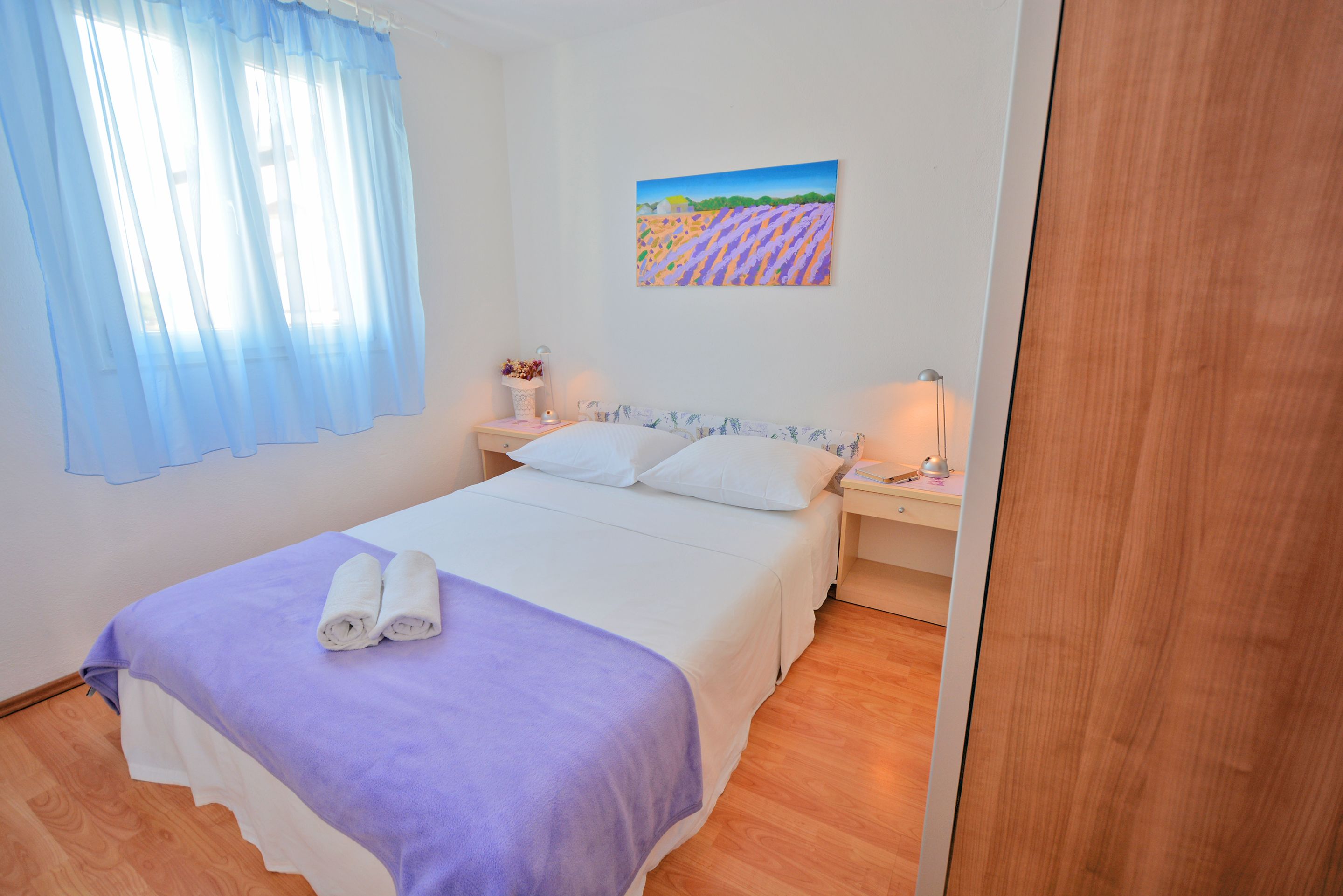 A neatly made bed with a purple blanket, a painting of a field on the wall, and a wooden floor.