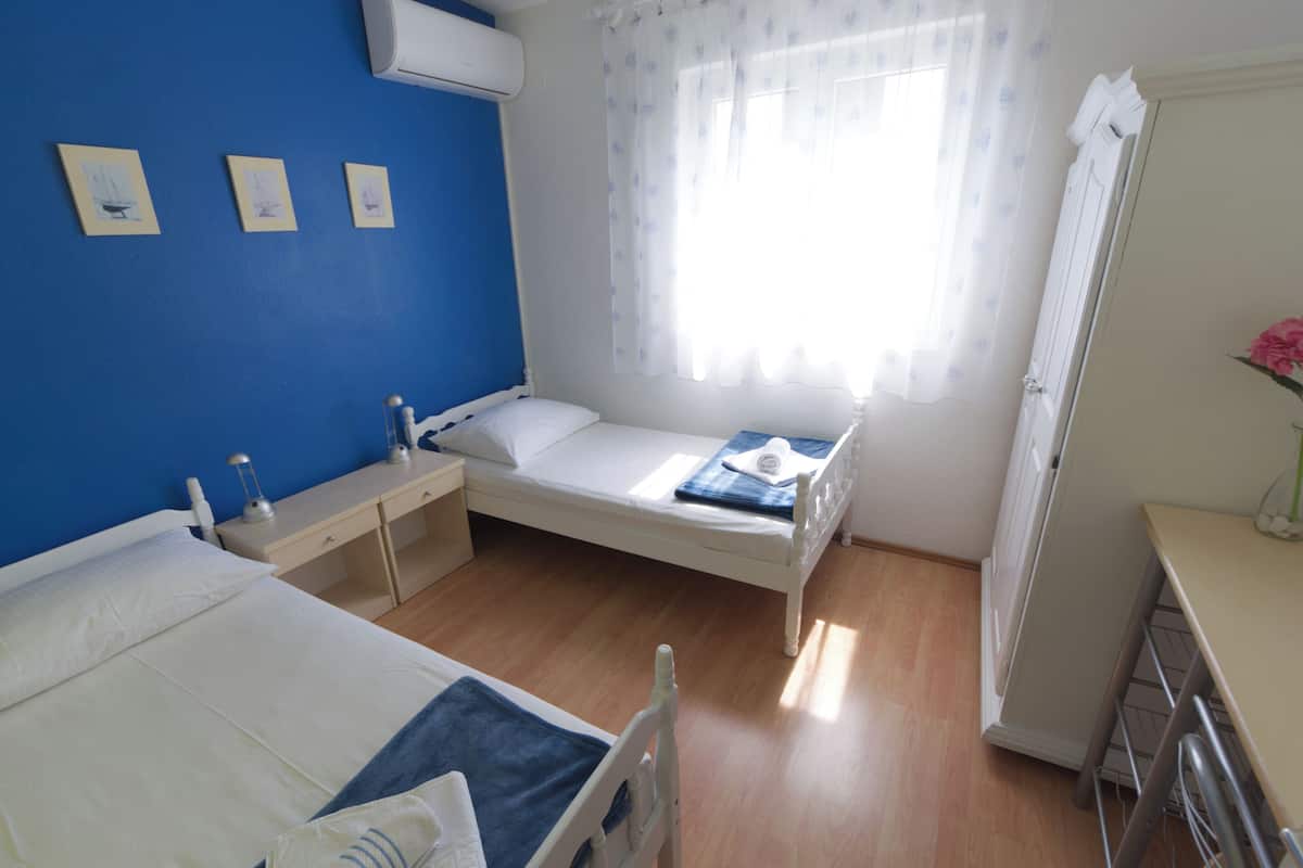 Een slaapkamer met een enkel bed, een nachtkastje, een klein bureau, een stoel, een raam met dunne gordijnen en een wandairconditioner.