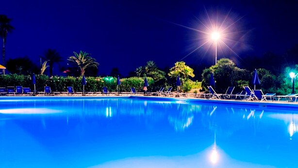 Ein Schwimmbecken bei Nacht mit klarem Wasser, umgeben von Liegestühlen und Sonnenschirmen, beleuchtet von hellem Kunstlicht.