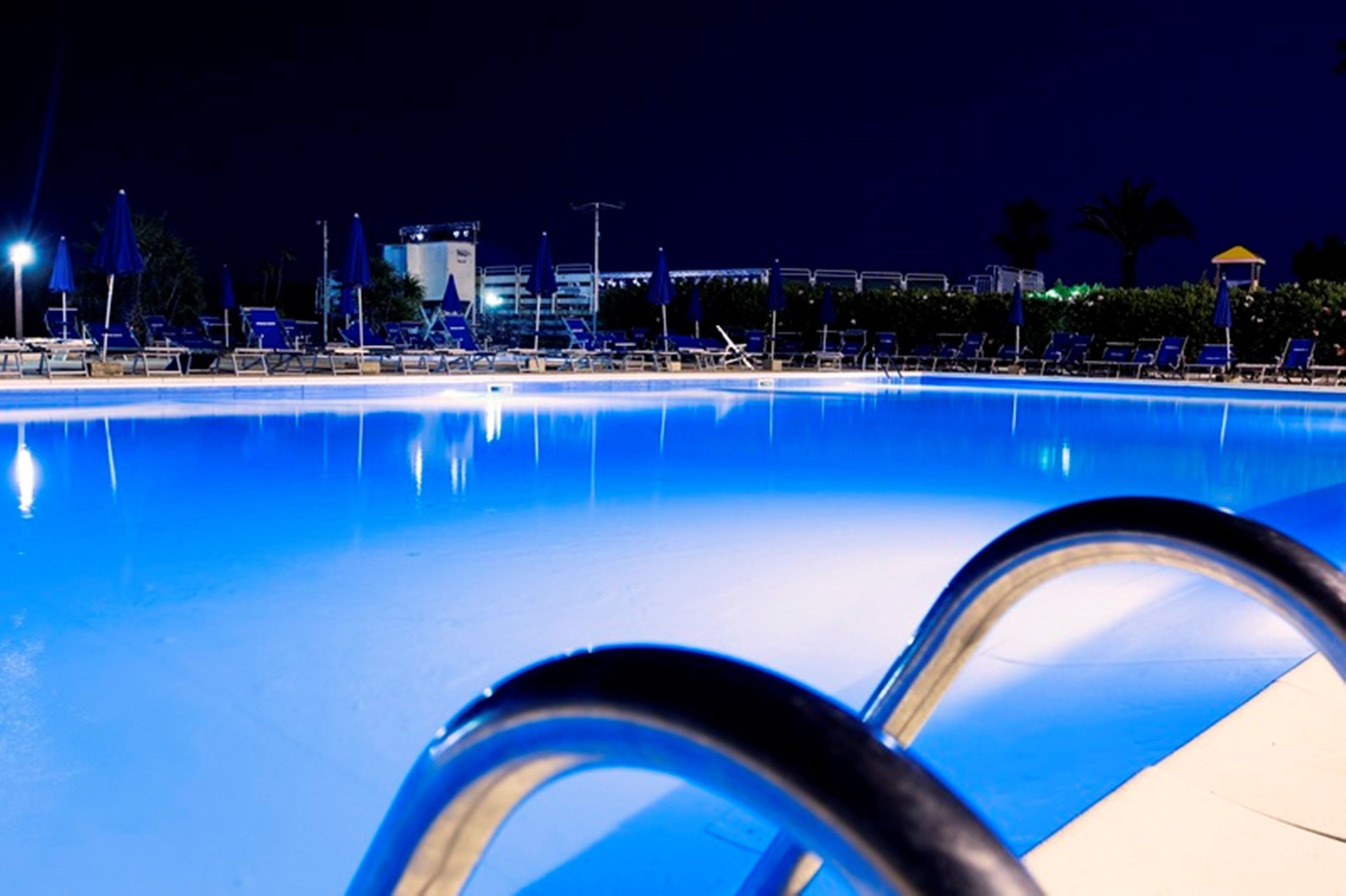 A nighttime pool area with lounge chairs and umbrellas, surrounded by buildings and palm trees.