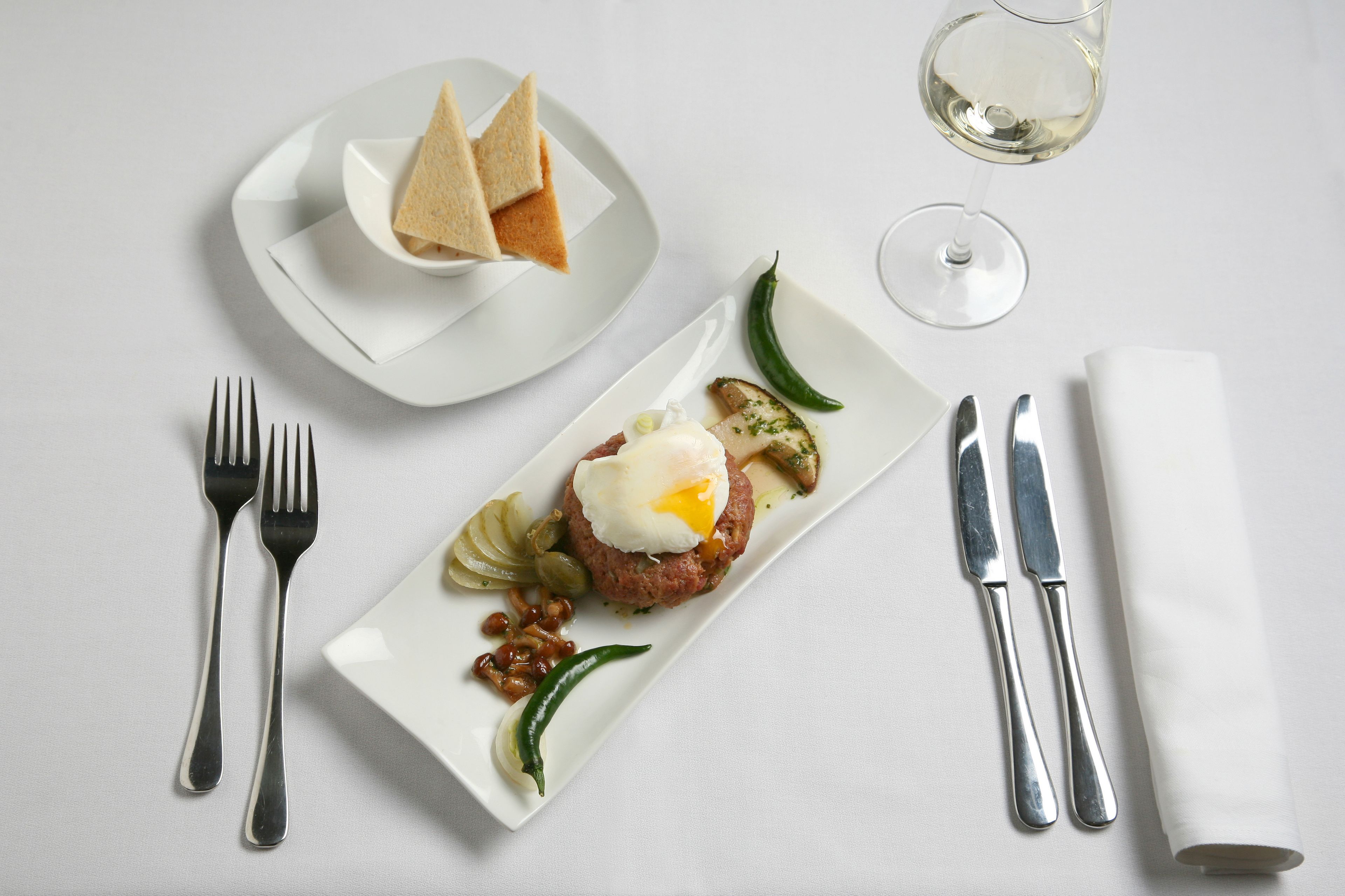 Ein ordentlich gedeckter Tisch mit einem Teller Essen, Besteck und einem Glas Weißwein.