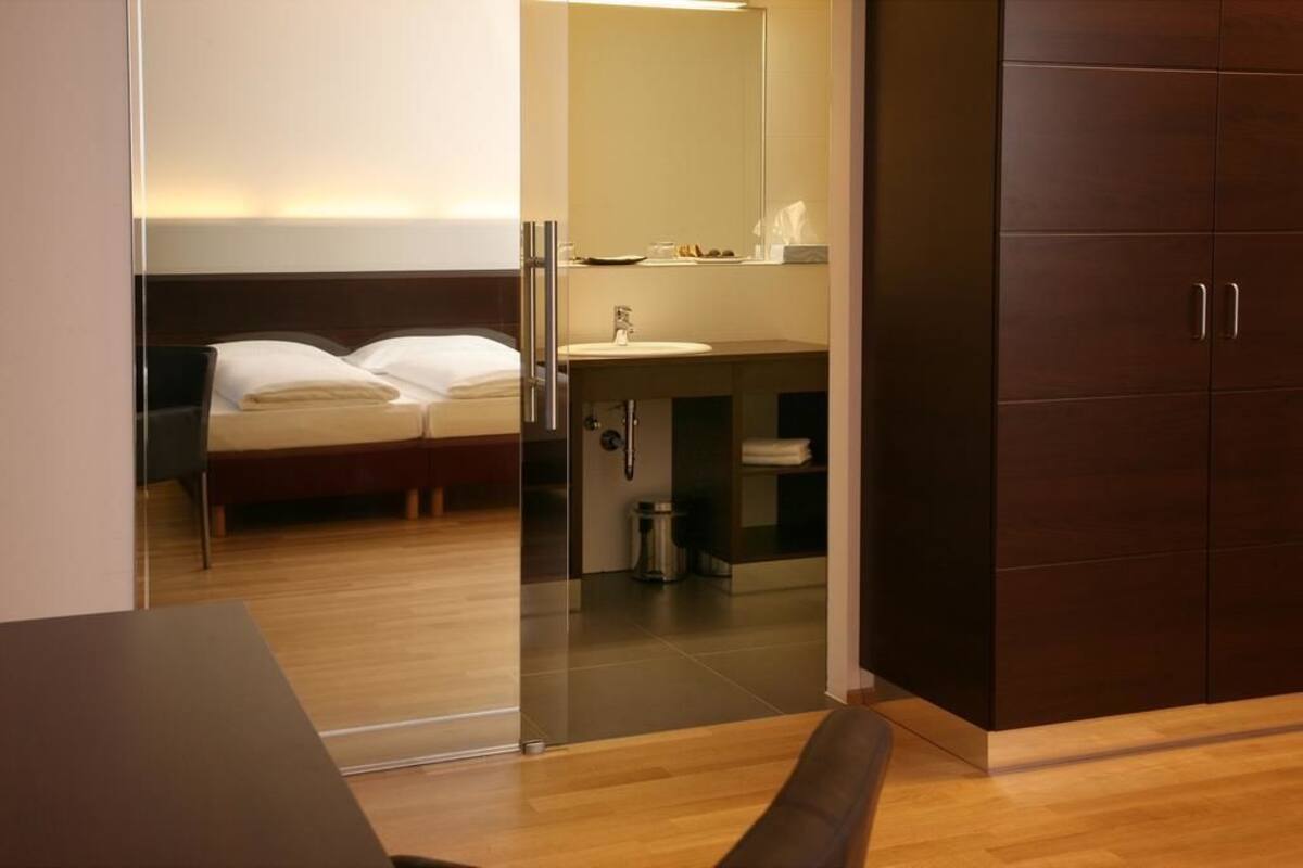 Quarto de hotel moderno com piso de madeira, cama com roupa de cama branca, banheiro com espelho, pia e porta-toalhas.