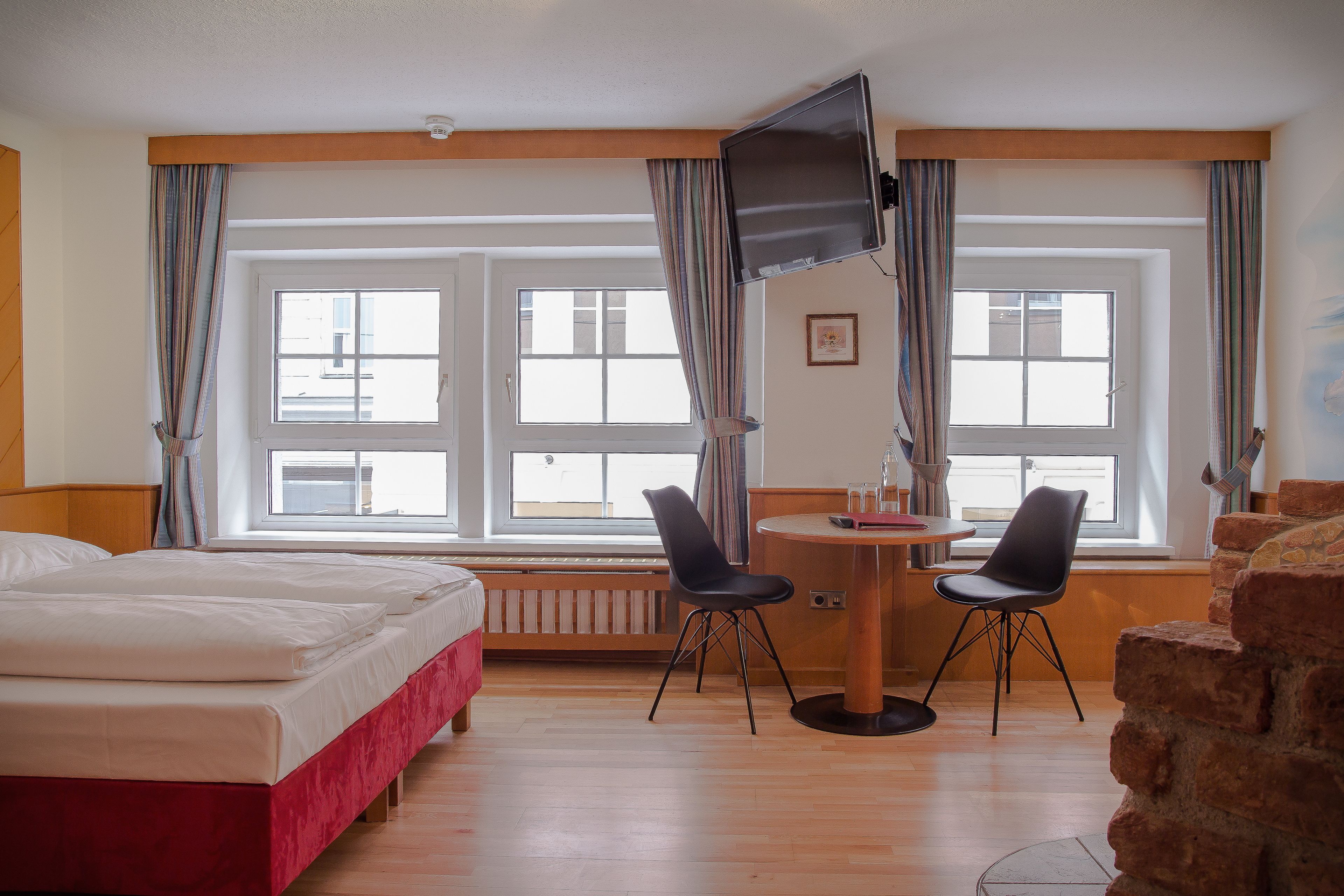 Habitación de hotel con una cama, dos sillas, una mesa, televisor y ventanas amplias.