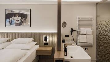 Una habitación de hotel moderna con una cama grande, una mesita de noche de madera, un tocador empotrado con espejo y perchero, y una obra de arte colgada en la pared.
