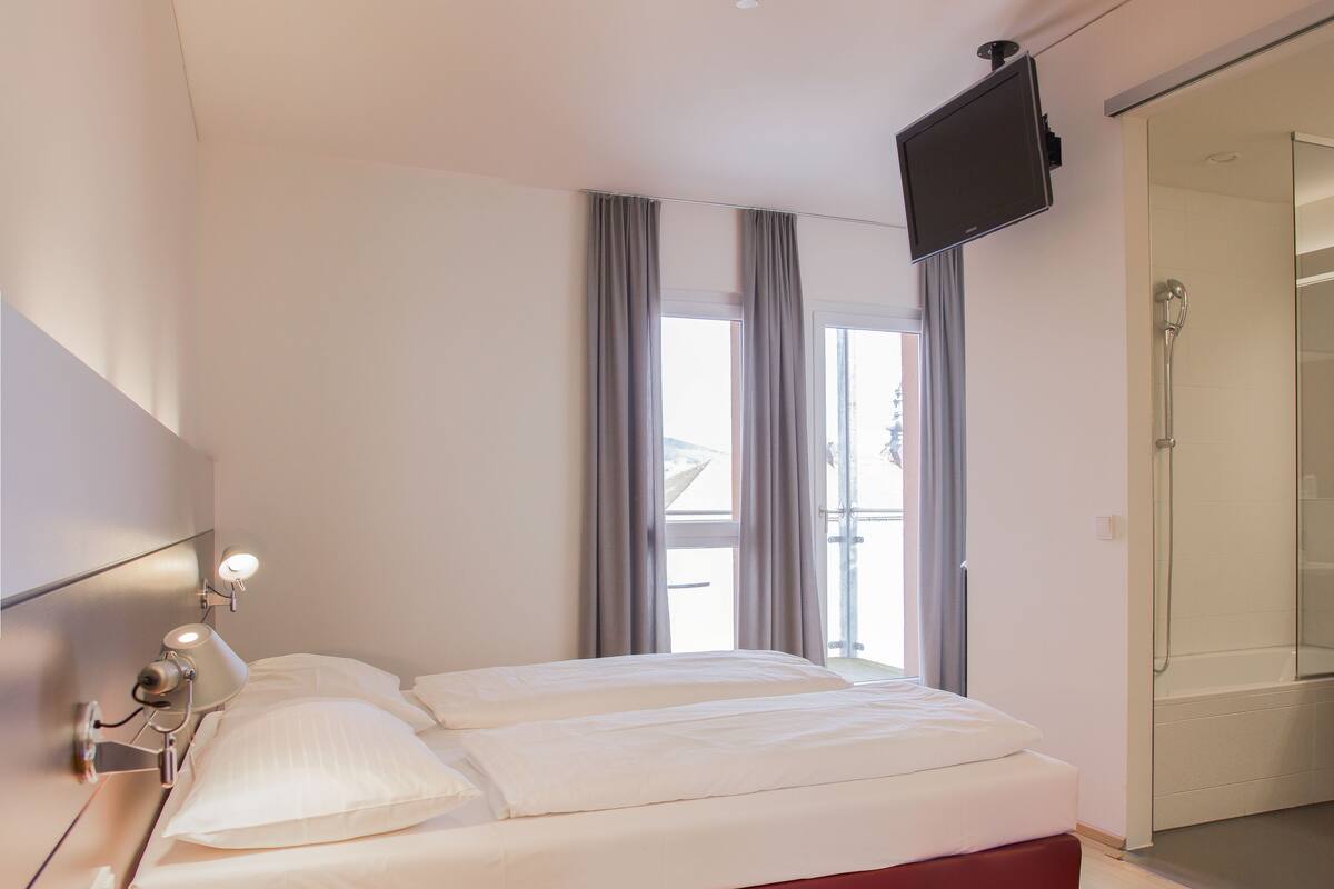 Quarto de hotel moderno com uma cama grande, mesa de cabeceira, televisão na parede e porta de vidro deslizante que dá para uma varanda.