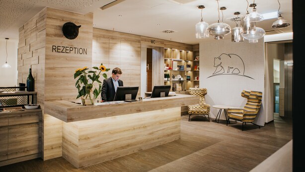 A reception area with a wooden reception desk, two computers, a reception counter, and a wall-mounted cat sculpture. - Hotel Schwarzer Bär (Linz)