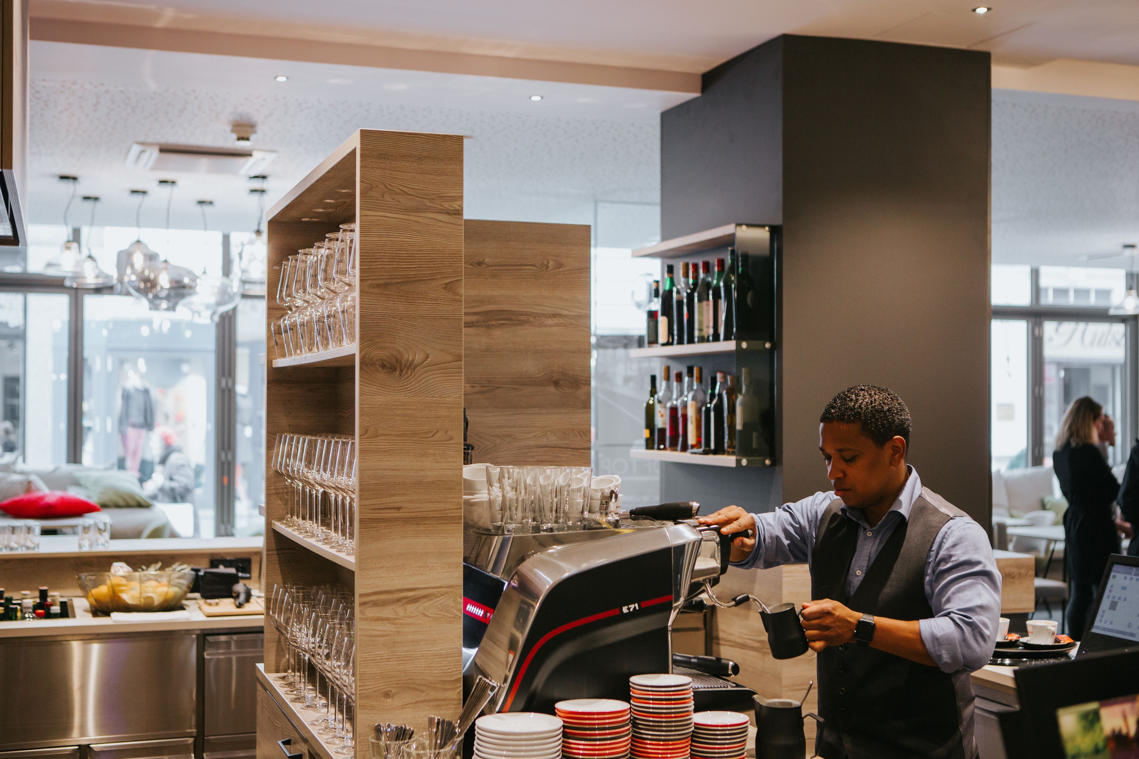 En barista laver en drik ved en disk med kaffemaskine, glas og en hylde med vinflasker i baggrunden.