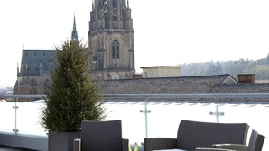A rooftop terrace with wicker furniture, a potted plant, and a view of a church spire. - Hotel Schwarzer Bär (Linz)