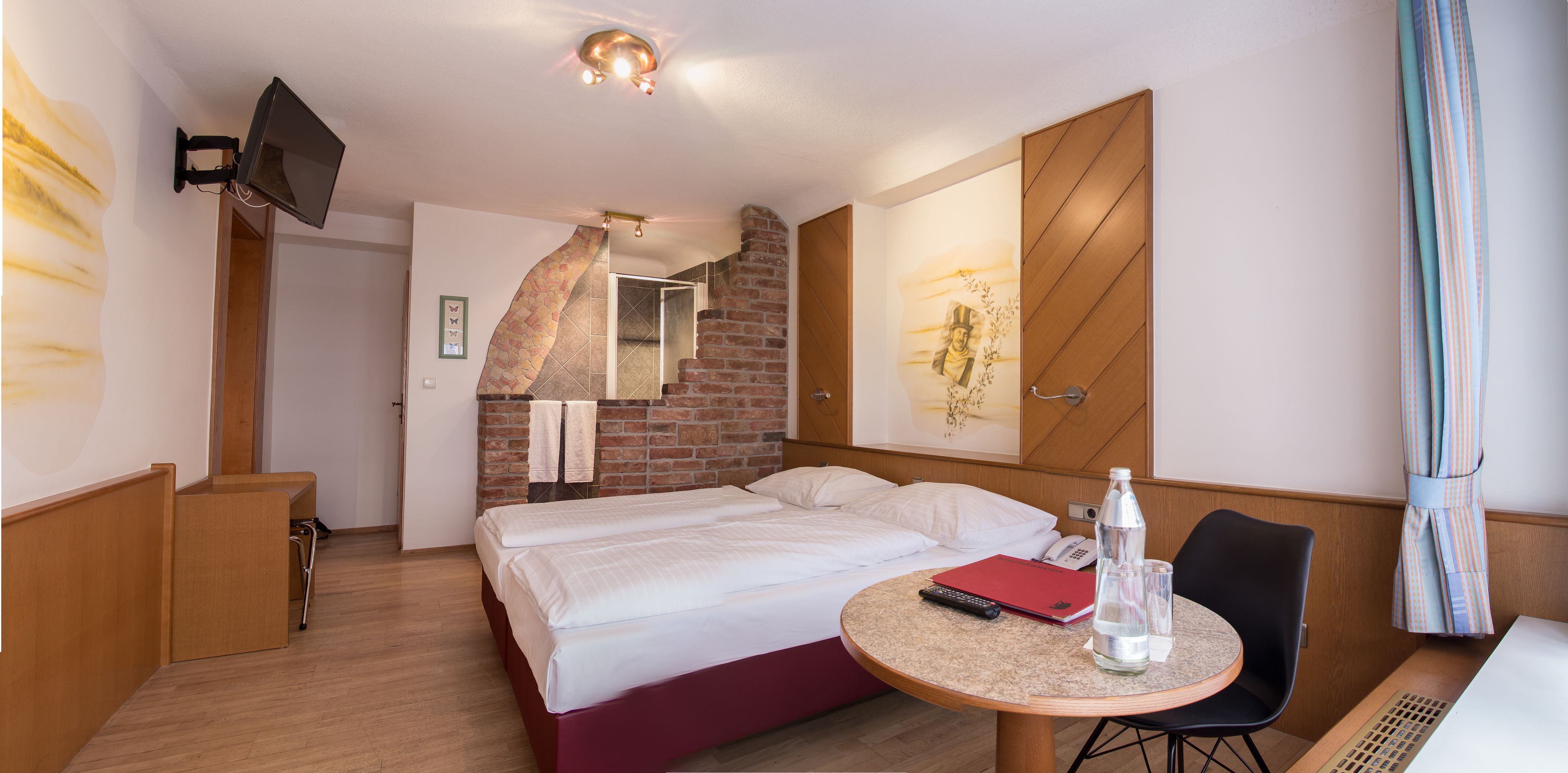 A hotel room with a bed, a round table with a chair, a TV mounted on the wall, and a brick wall design.