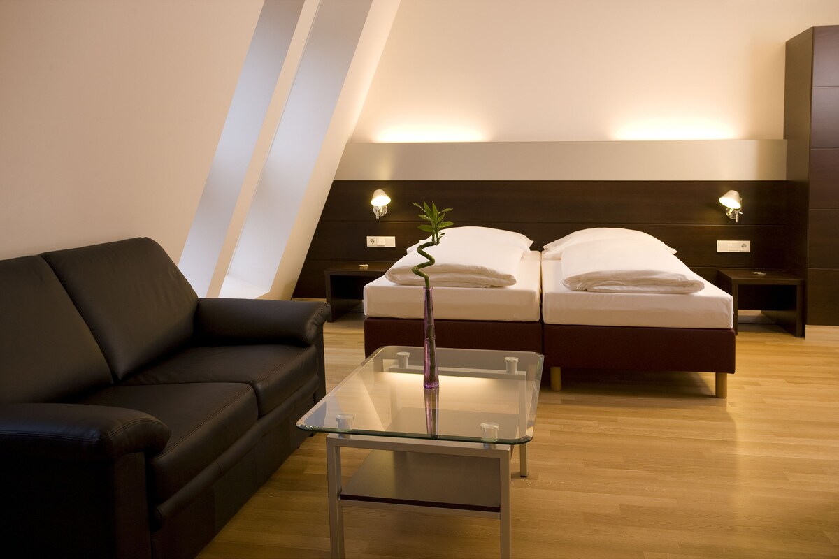 A modern hotel room with a glass coffee table, a black sofa, a bed with white linens, and two wall-mounted nightstands.