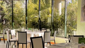 Ein Restaurant mit runden Tischen, das von großen Fenstern gesäumt ist und einen Blick auf üppiges Grün bietet.