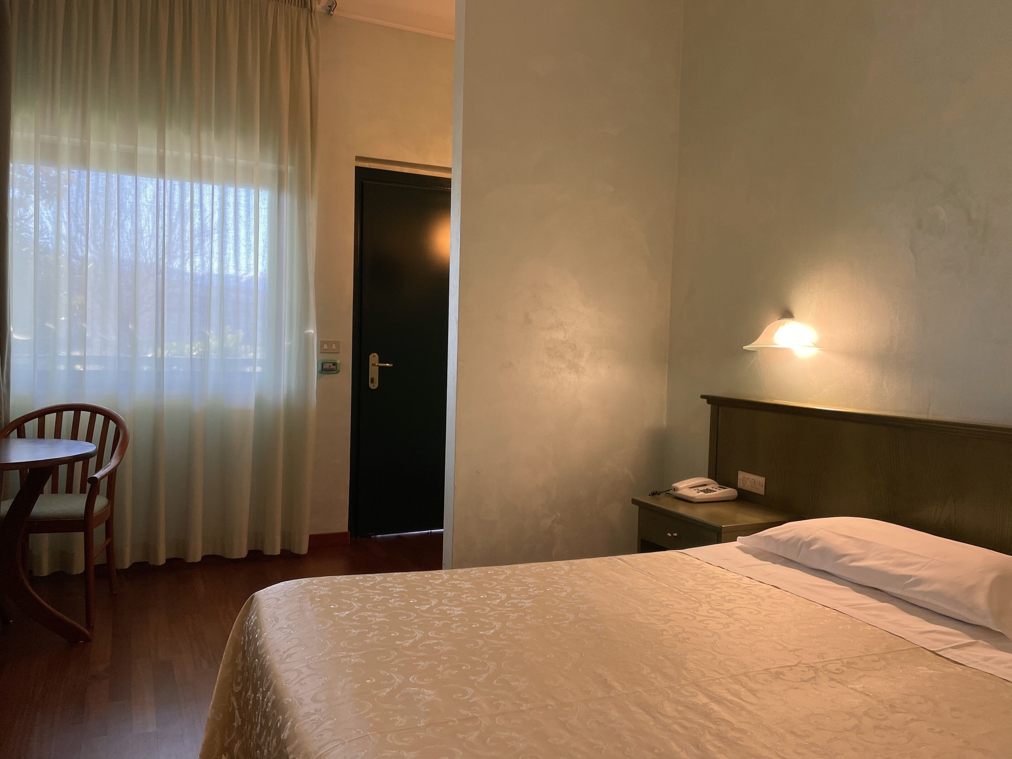 A hotel room with a bed, a chair, a desk with a phone, and a door.