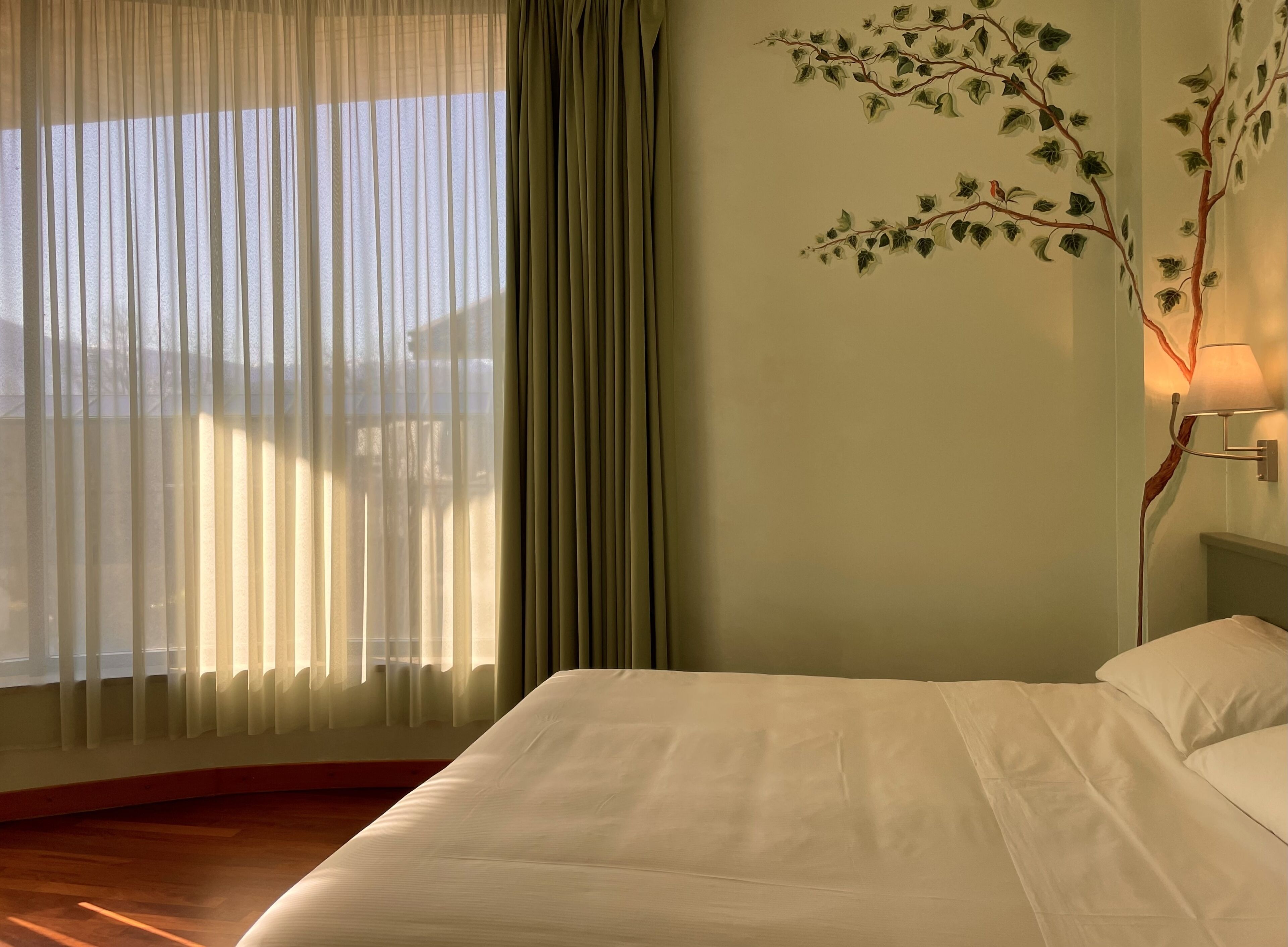 A hotel room with a bed, a window with sheer curtains, a wall-mounted lamp, and a view of a building outside.