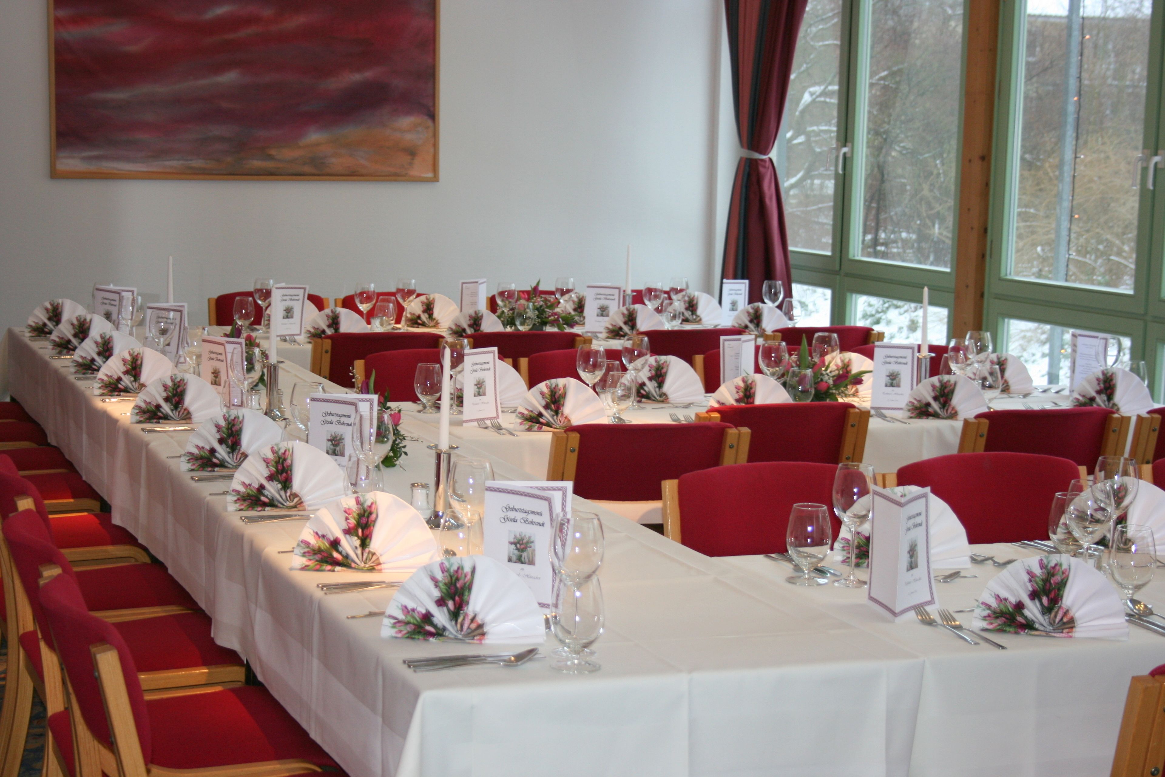 Un salón de banquetes con largas mesas dispuestas para un evento formal, con manteles blancos, sillas rojas y centros de mesa florales.