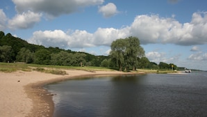 A riverside landscape with a sandy beach, trees, and a boat in the distance. - Parkhotel Hitzacker (Hitzacker)