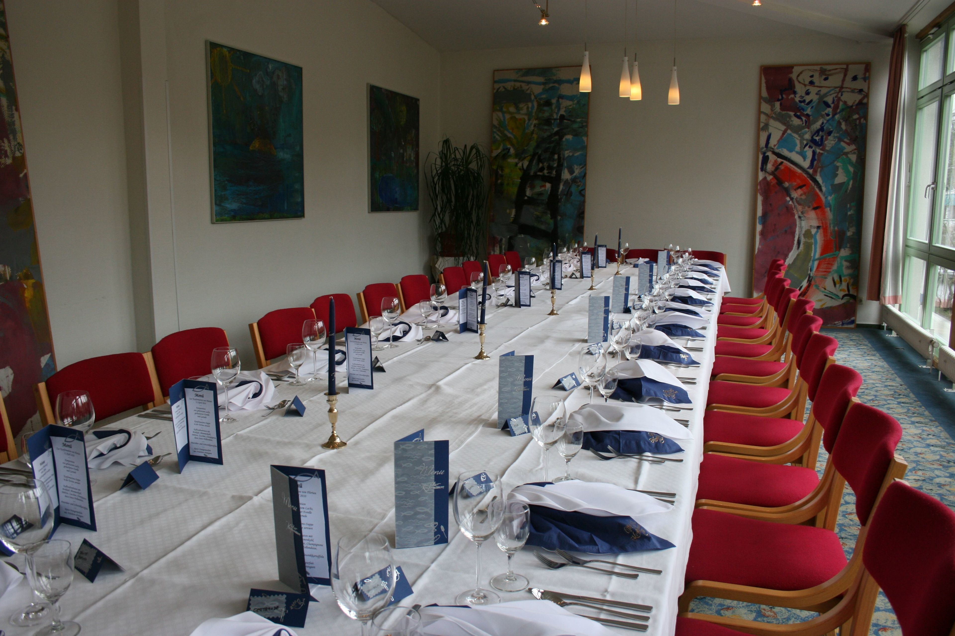 Una larga mesa de comedor con sillas rojas, manteles blancos y atril para menús azules.