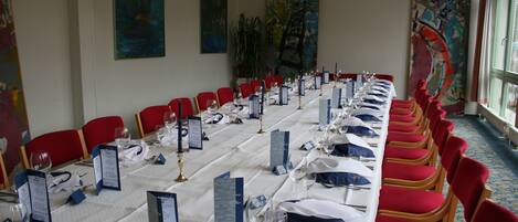 Una larga mesa de comedor con sillas rojas, manteles blancos y atril para menús azules.