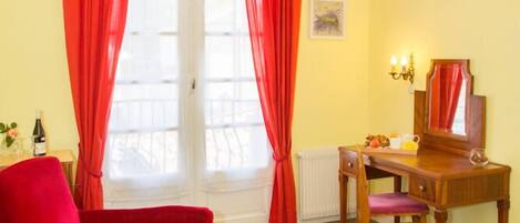 A room with a red chair, a wooden desk, a small table with a vase, and a chandelier.