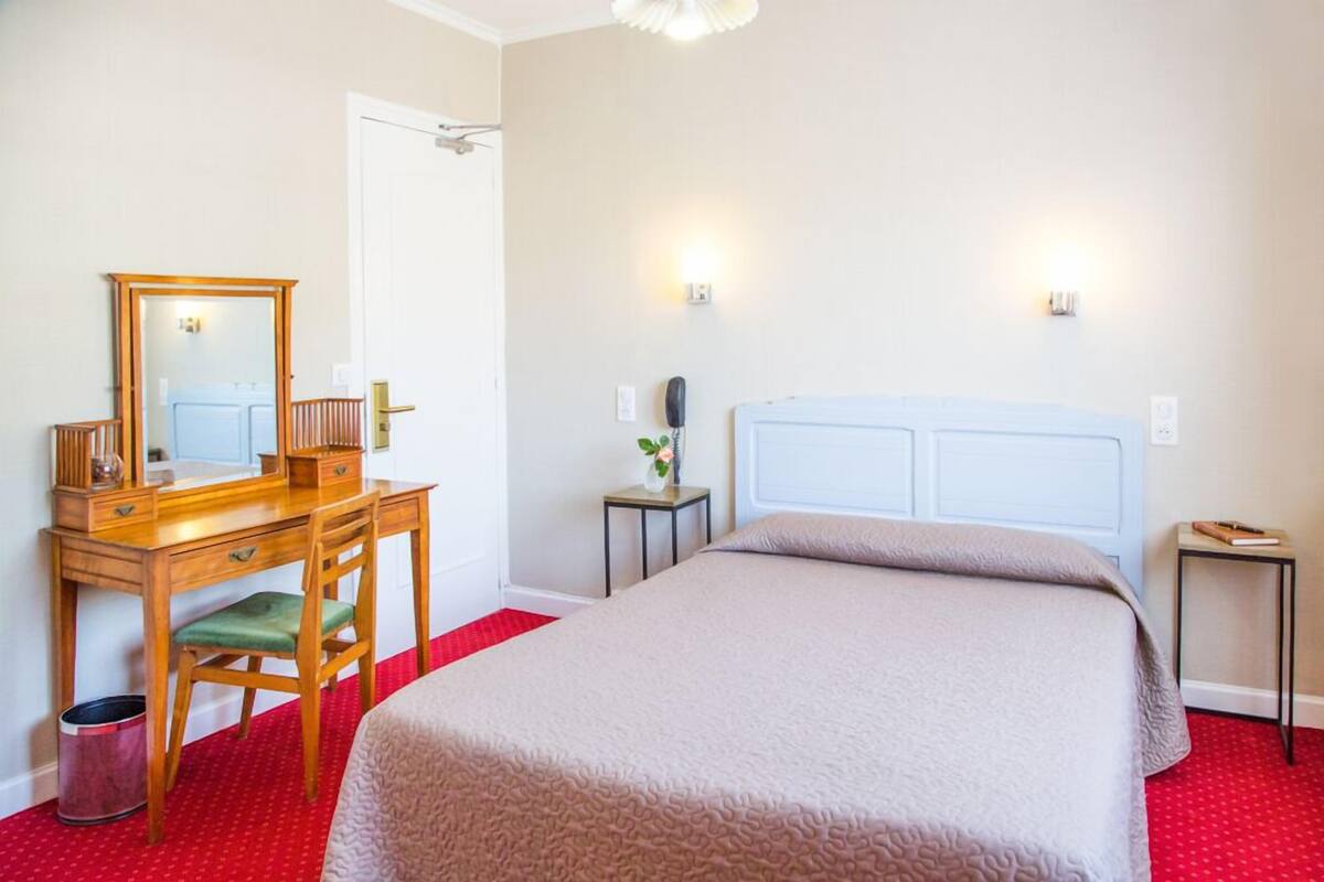 Una habitación de hotel con una cama, un escritorio con silla, un espejo, una mesita de noche y una alfombra roja.