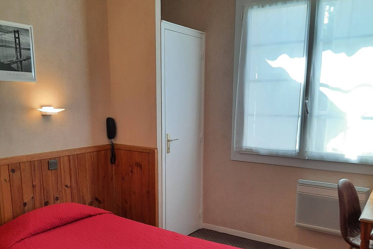 Una habitación con cama, escritorio, silla, teléfono, radiador y ventana.