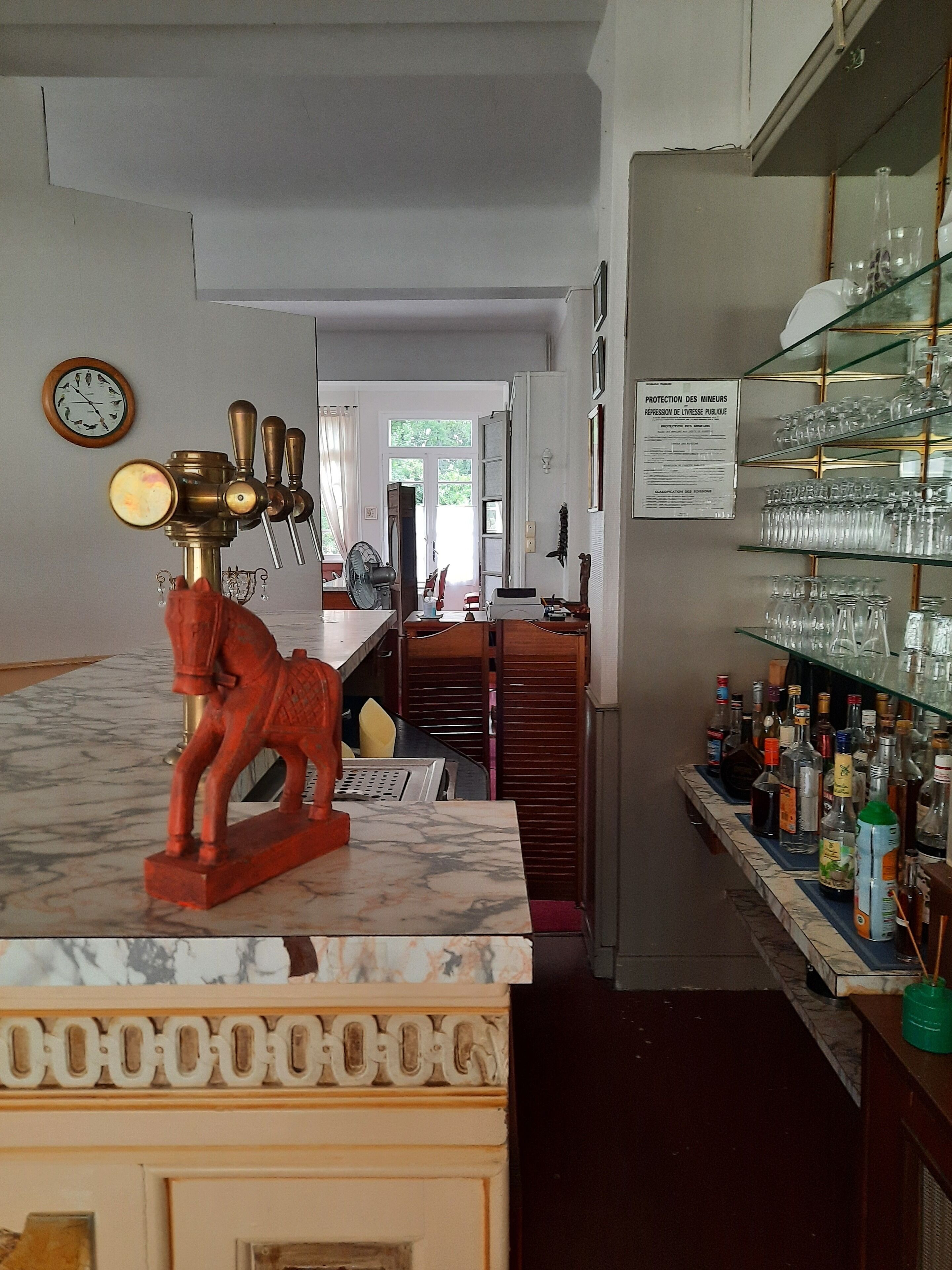 Eine Bar mit Marmorplatte, einer roten Skulptur eines Pferdes, einer Uhr und Regalen mit Gläsern und Flaschen.