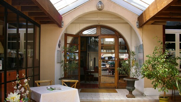 Une terrasse extérieure avec une table et des chaises, un pot de fleurs et une arche en bois menant à un bâtiment.