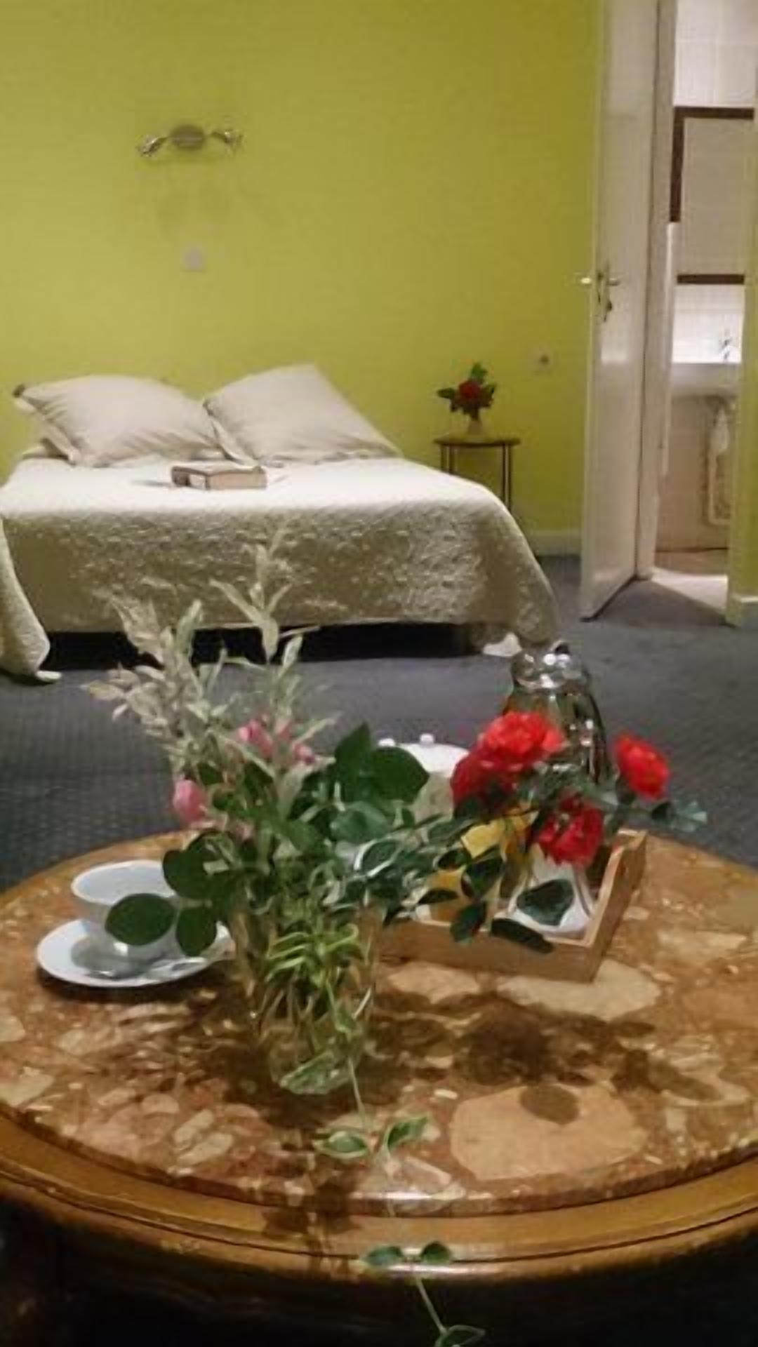 A room with a bed, a small table with a vase of flowers, and a door leading to another room.