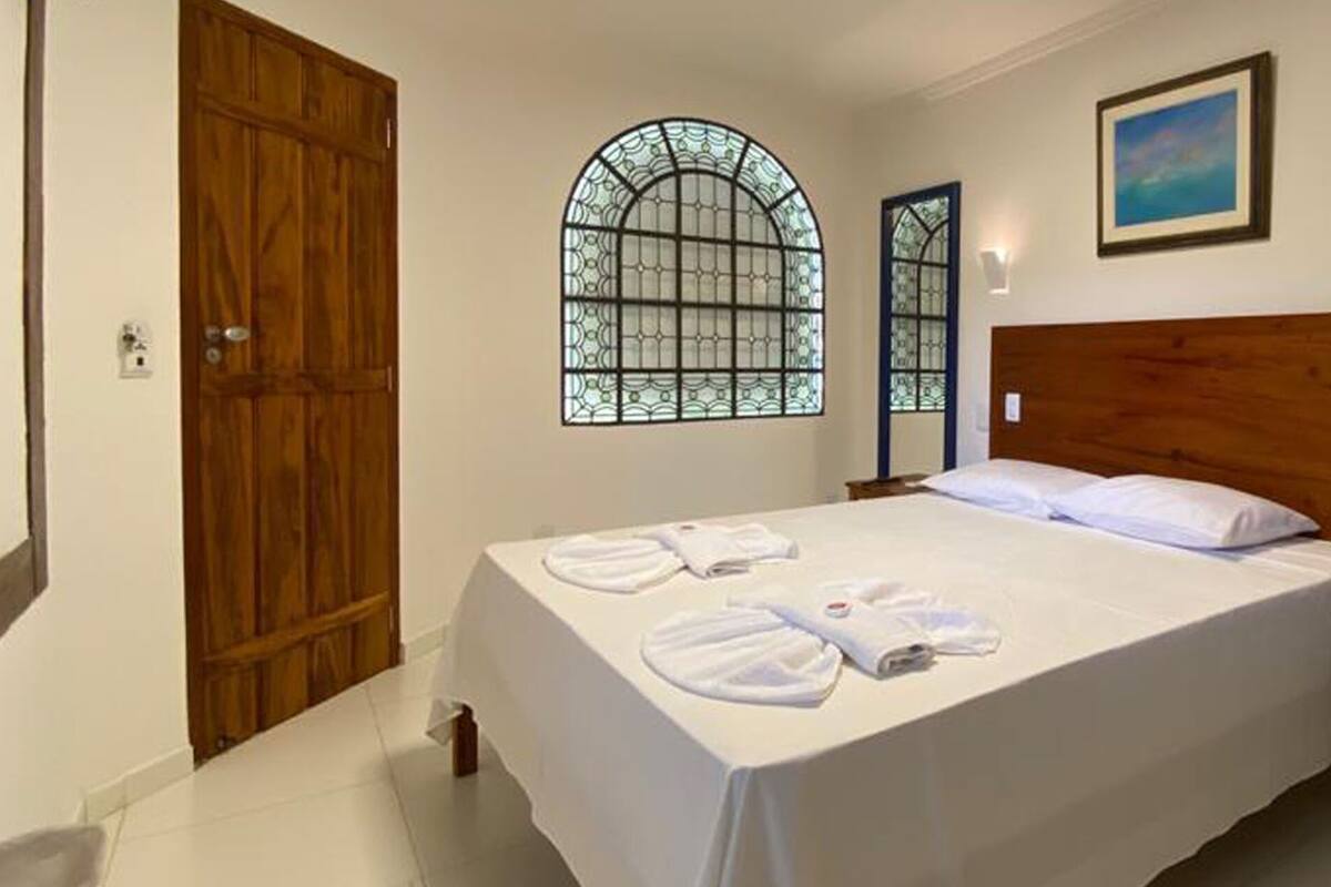 A hotel room with a large bed, two windows with stained glass, a wooden headboard, a bedside table, and a radiator.