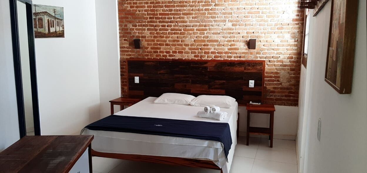 A bedroom with a brick wall, a wooden headboard, a bed with white linens, two bedside tables, and a small wooden table.