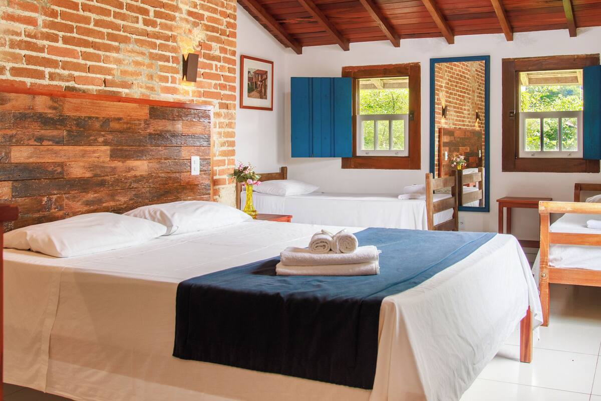 A hotel room with a brick wall, wooden ceiling, two beds, a wooden bench, and a window with blue shutters.