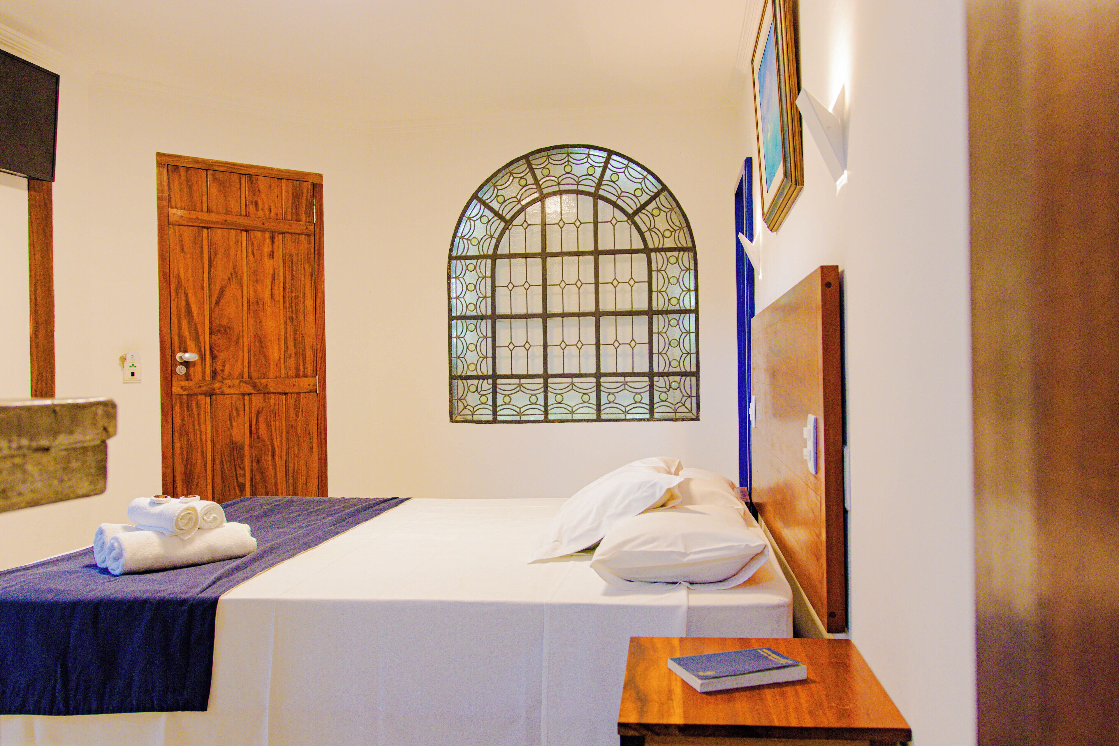 A hotel room with a bed, a wooden door, a stained glass window, and a small table with a book.
