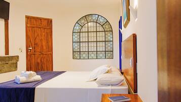 A hotel room with a bed, a wooden door, a stained glass window, and a small table with a book.