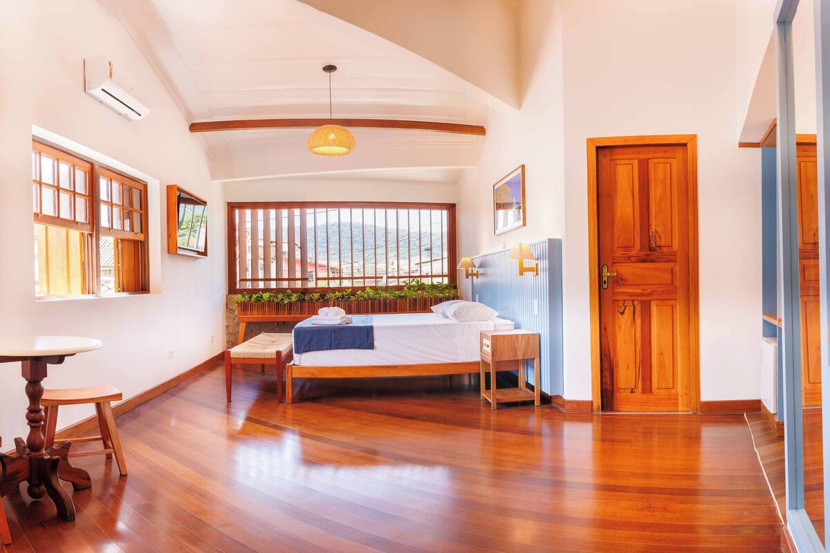 A bedroom with a wooden bed, a small table, a chair, a window with a view, and a wooden door.