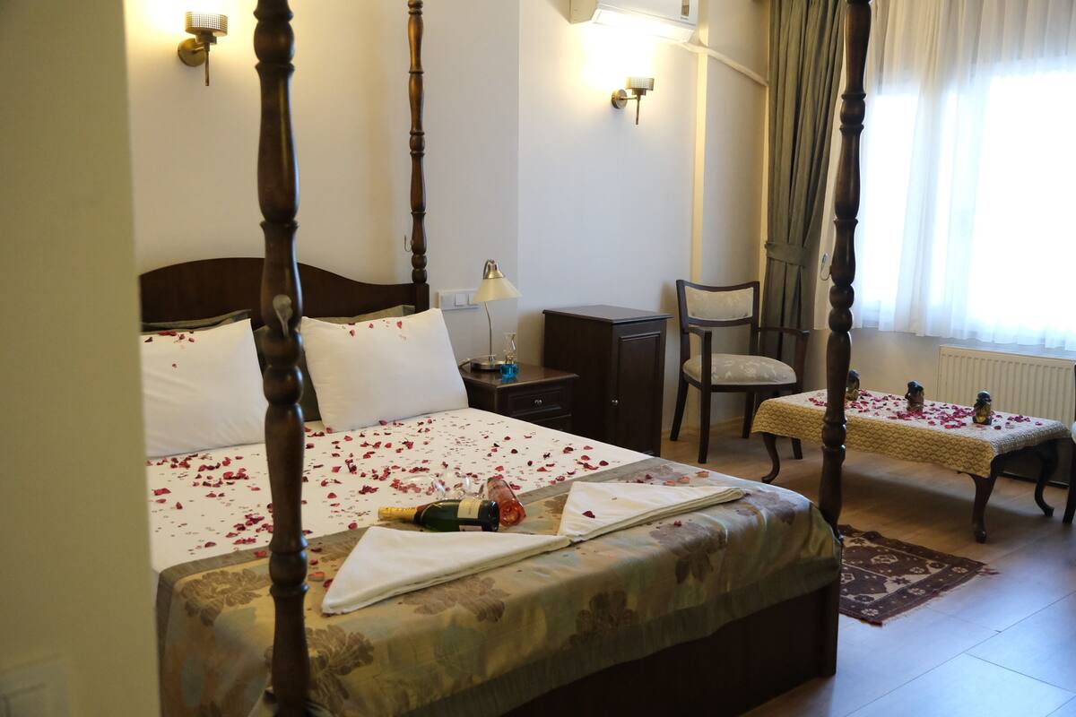 A four-poster bed with rose petals, a bedside table with a lamp, a chair, and a small table with a tablecloth and candles.