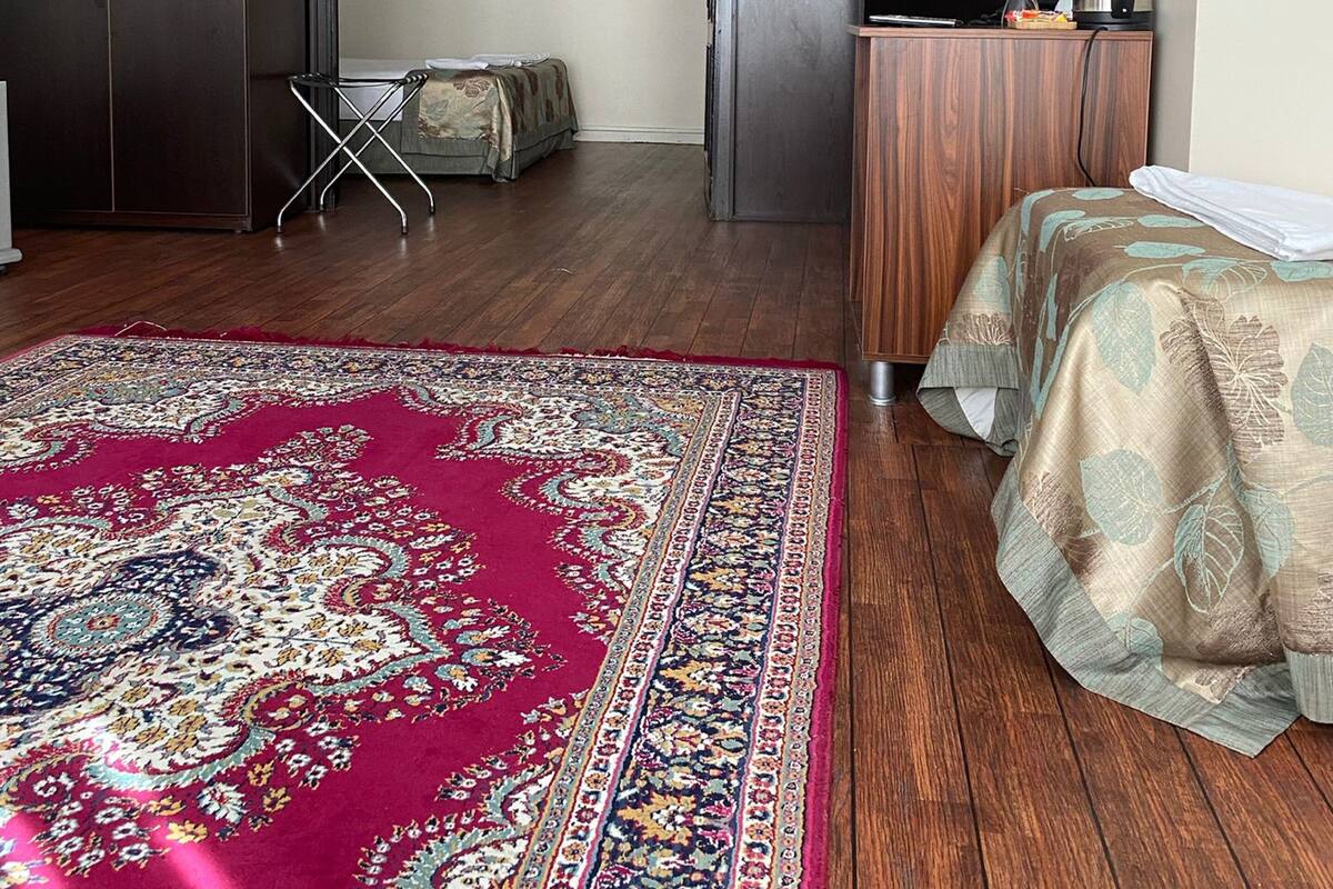 A room with a large red patterned rug, a bed with a green and beige bedspread, a wooden desk with a kettle, and a wardrobe.