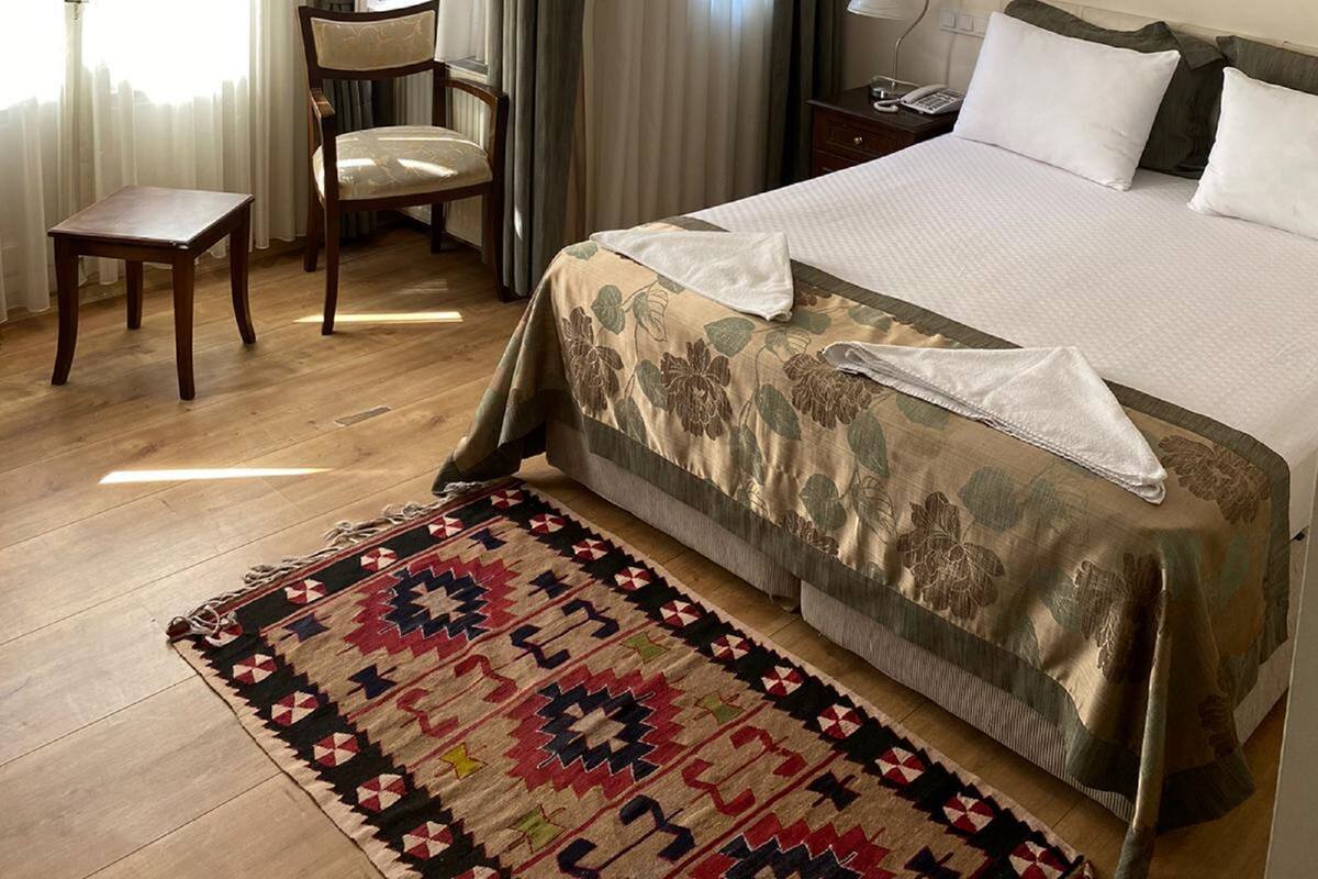 A hotel room with a bed, a desk, a chair, a lamp, and a patterned rug.