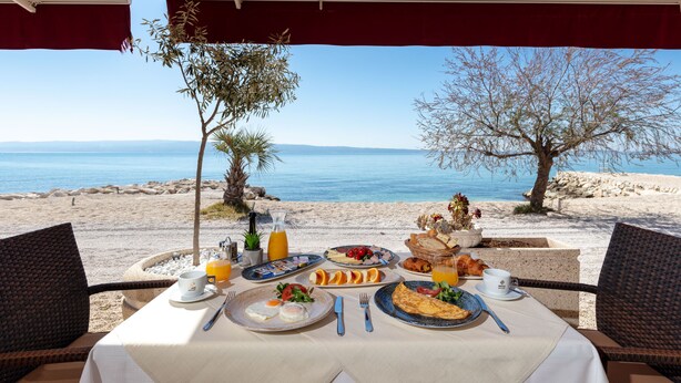 Une table dressée pour le petit-déjeuner, avec vue sur la mer, comprenant des assiettes de nourriture, des couverts et un verre de jus d’orange.
