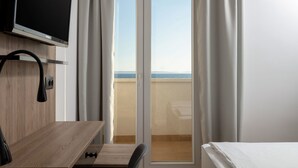 Une chambre d’hôtel avec un bureau en bois, un téléviseur à écran plat fixé au mur, un lit et une vue sur la mer, accessible par une porte-fenêtre coulissante.