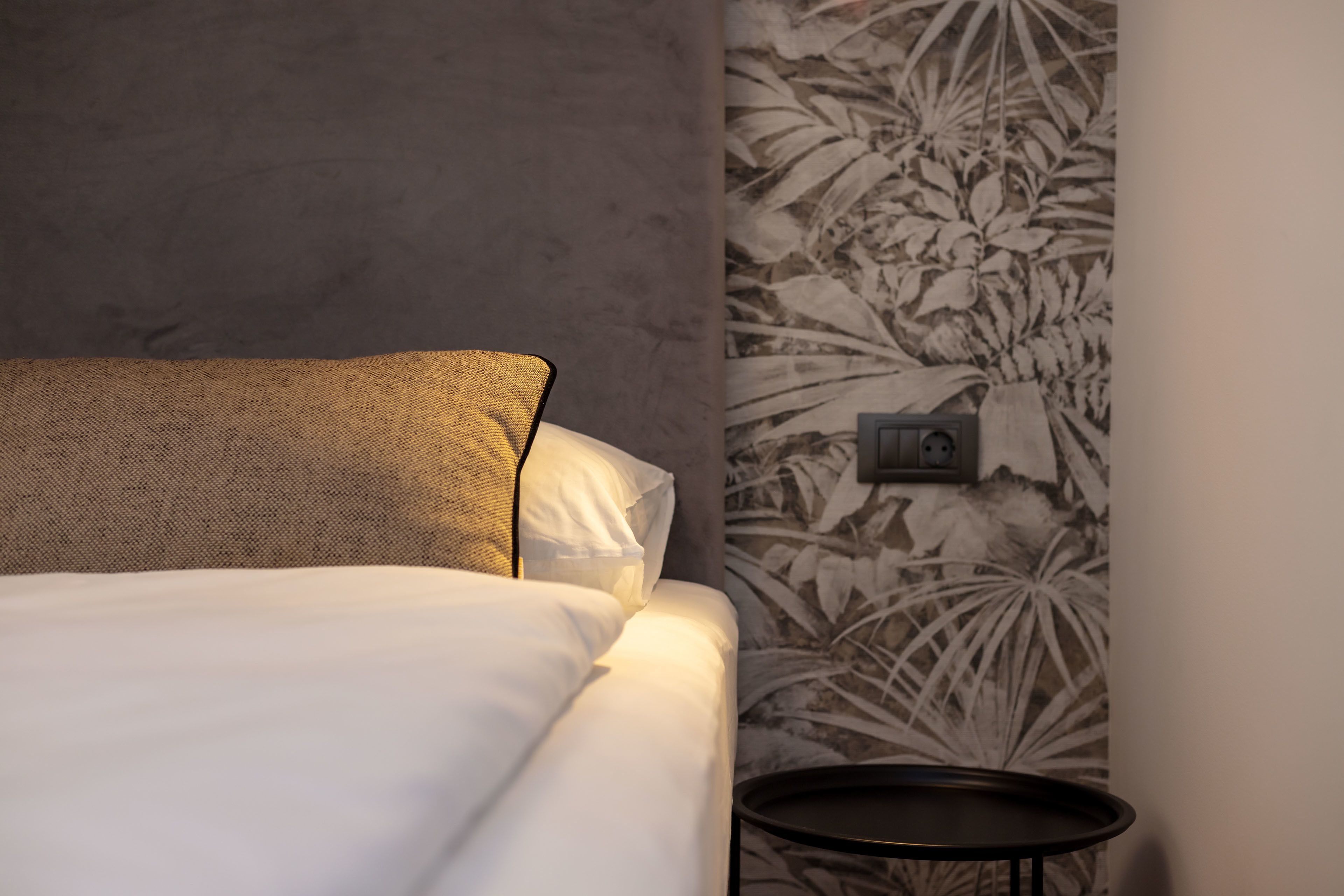 Un lit bien fait, avec du linge de lit blanc et un coussin à motifs, une petite table ronde à côté, et un mur recouvert d’un papier peint à motifs floraux.