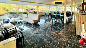 Ein großzügiger Innenbereich mit hoher Decke, eine Sitzecke mit Sofas und Sesseln, ein Essbereich mit Tischen und Stühlen und Blick auf Grünflächen draußen.