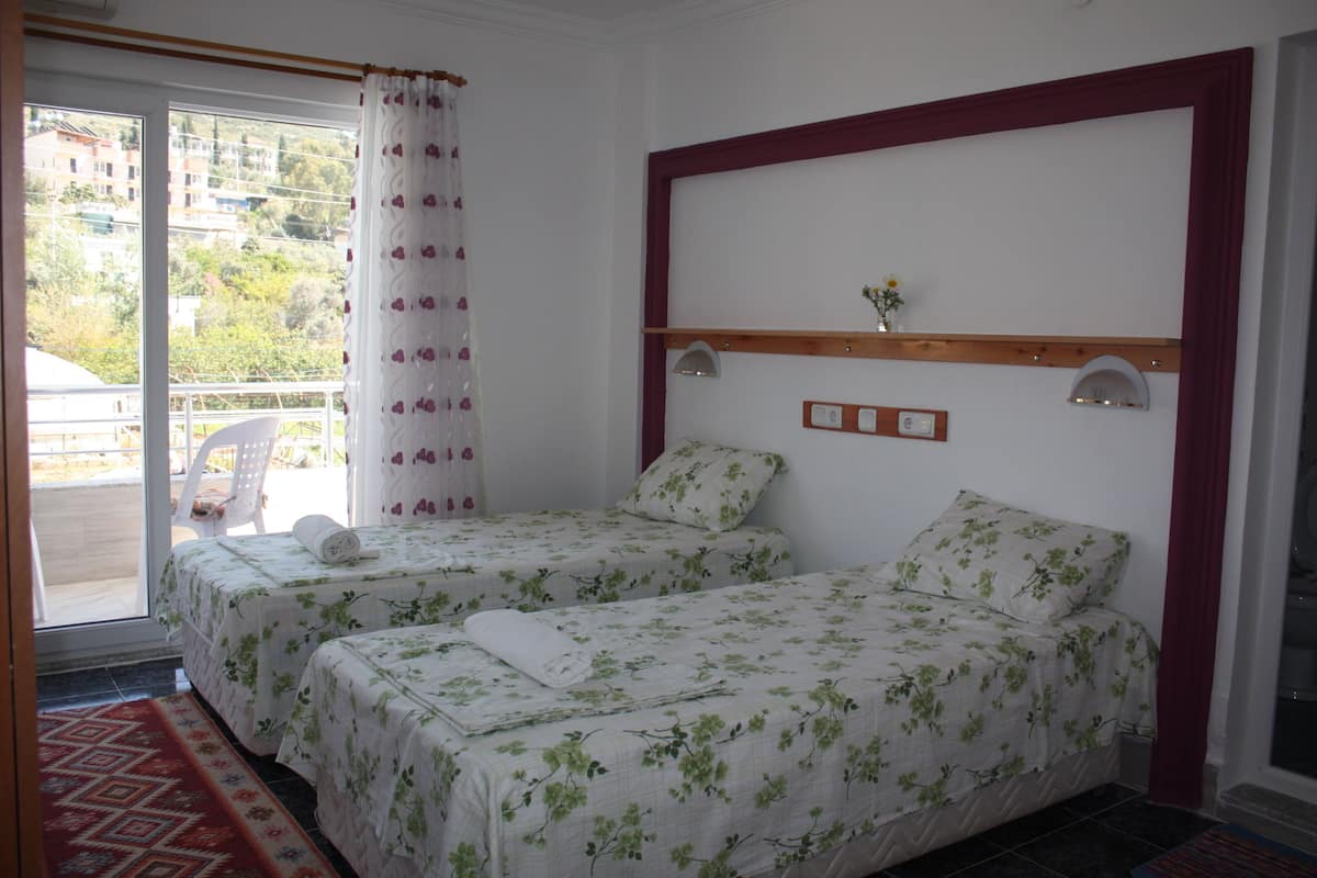 A room with two beds, a window with a view of buildings, a wooden headboard, and a small shelf with a vase.
