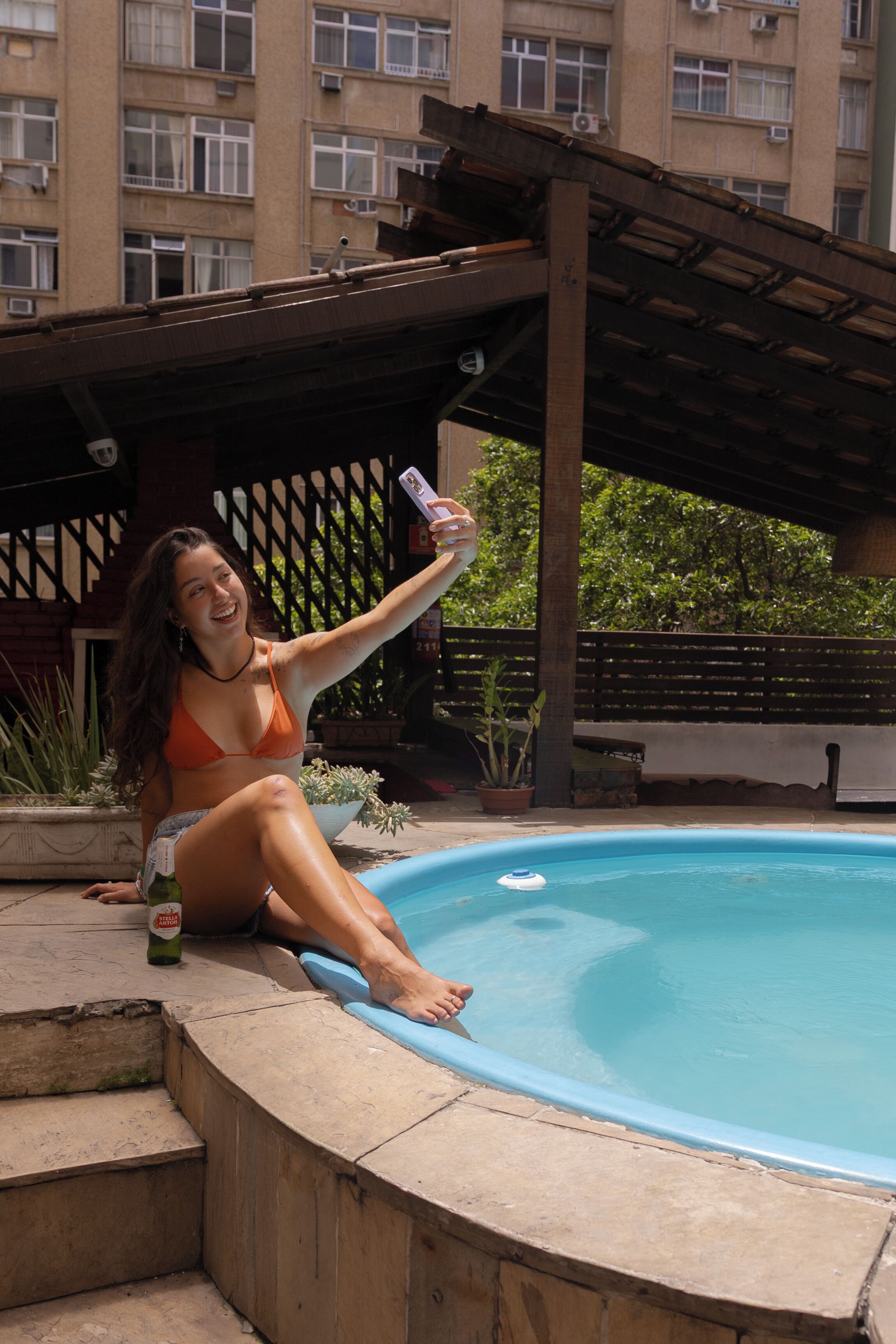 Una persona sentada junto a una piscina bajo una pérgola.